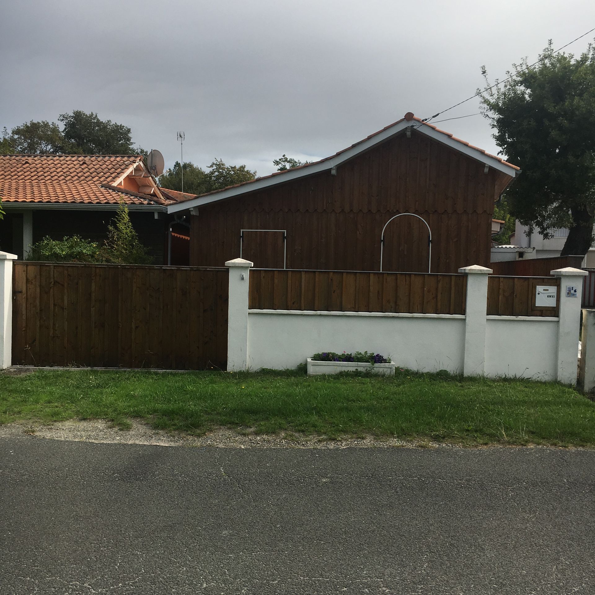 Maison brune derrière une clôture en bois brun, mur blanc, herbe verte, ciel couvert.