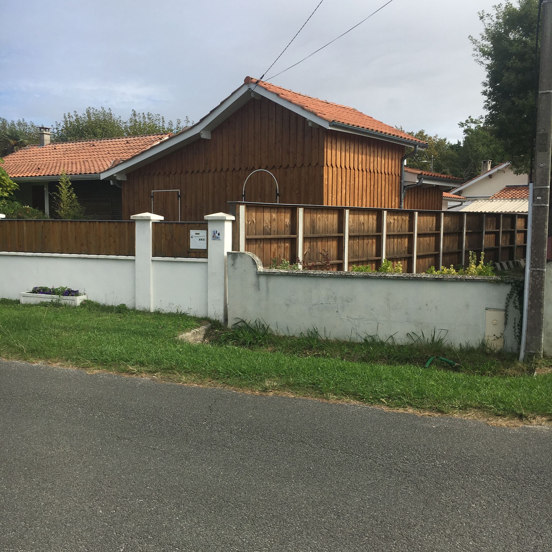 Une maison en bois brun avec un toit de tuiles rouges, derrière un mur blanc et une clôture en bois, vue depuis la route.