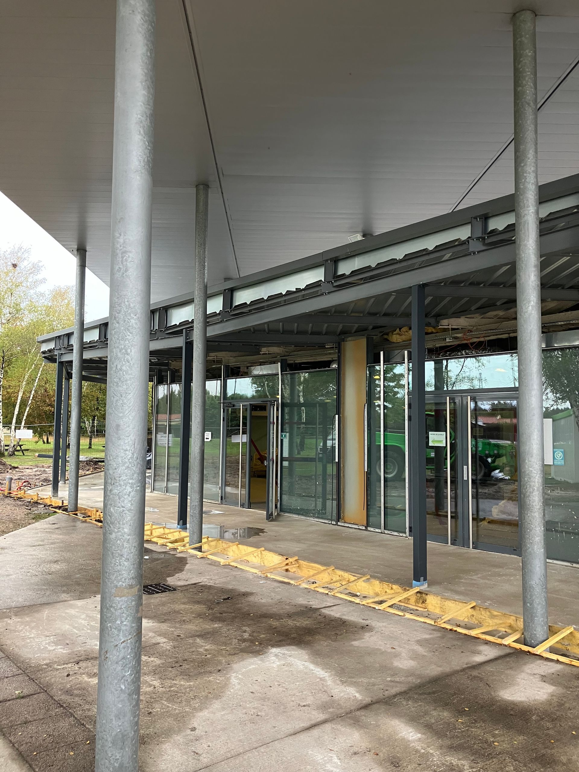 Entrée couverte avec portes vitrées, soutenue par des poteaux métalliques, avec des barrières de chantier au sol.