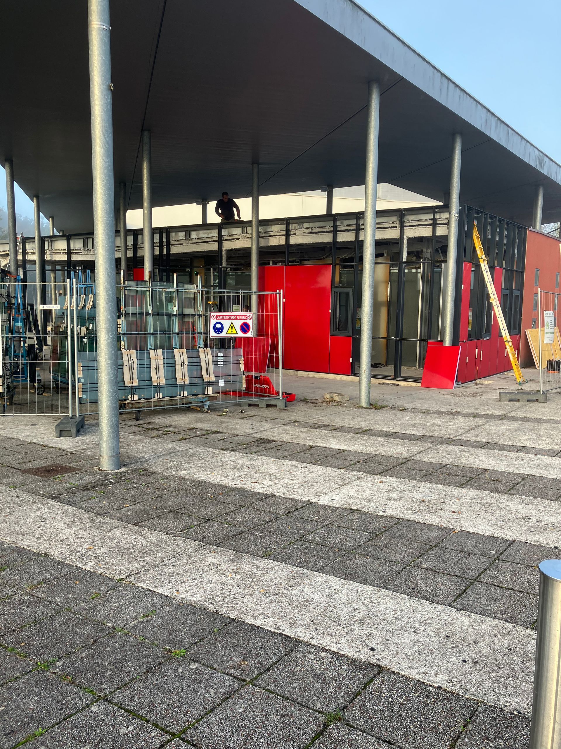 Bâtiment en construction, panneaux rouges, sol pavé gris et blanc, supports métalliques et échafaudages.