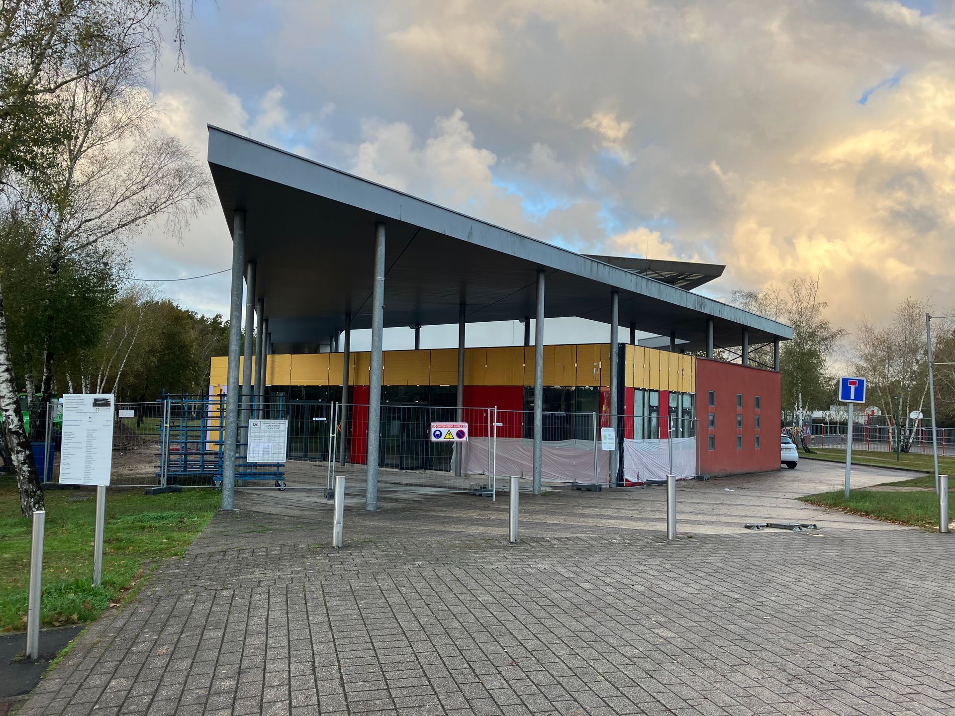 Bâtiment moderne au toit incliné, soutenu par des poteaux. Parties rouges, jaunes et en briques. Zone pavée au premier plan.