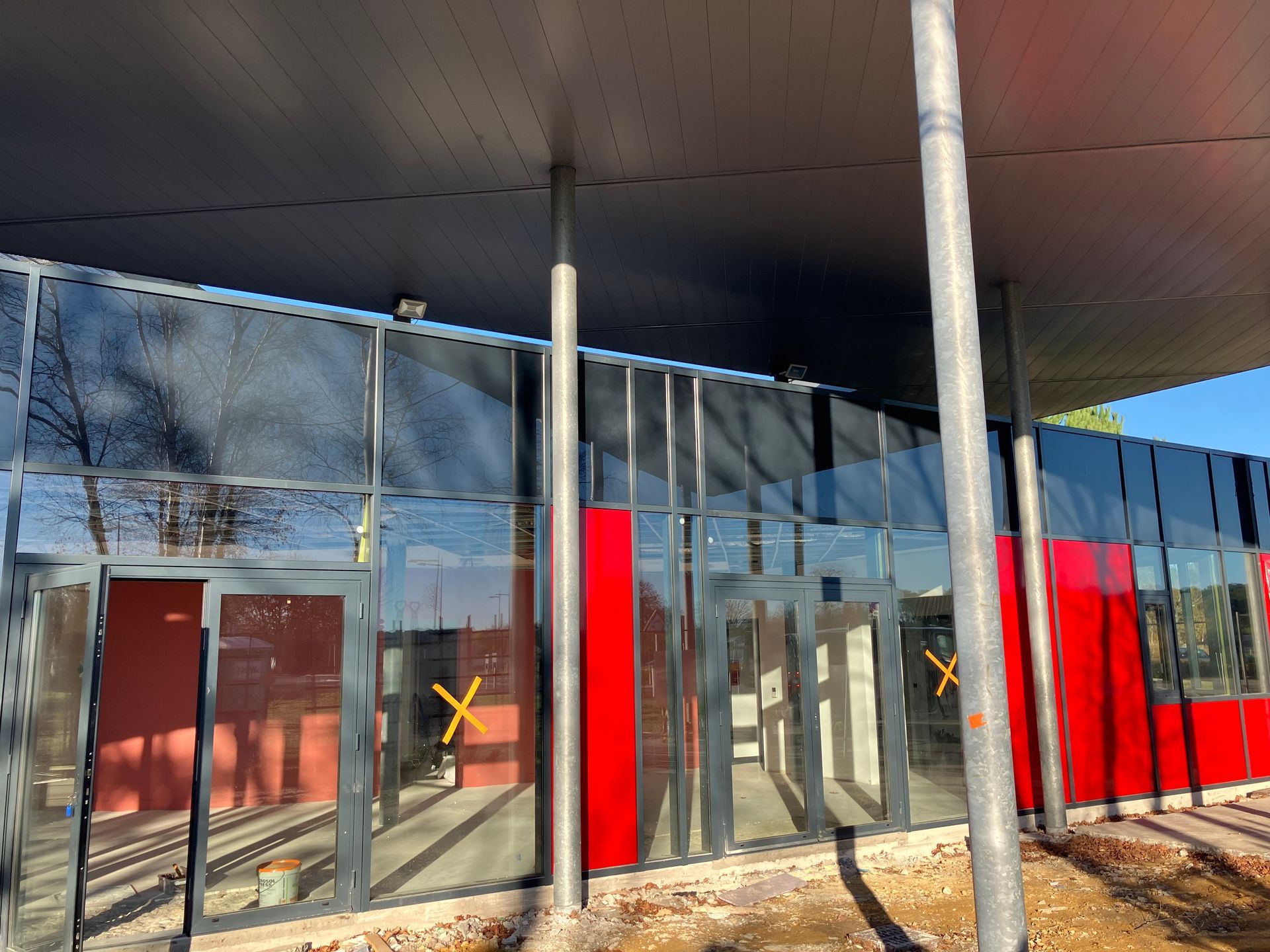 Bâtiment aux murs de verre, orné de touches de rouge et d'un toit sombre, soutenu par des colonnes métalliques.