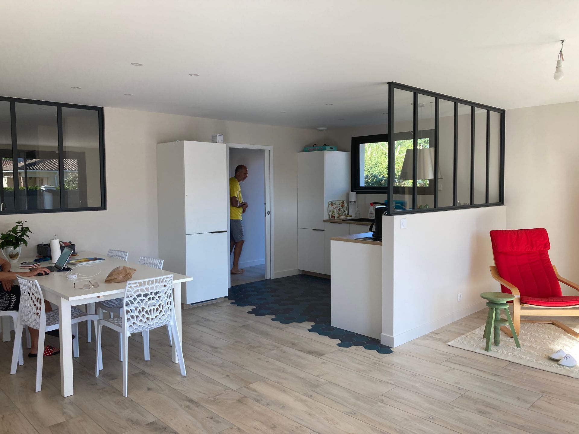 Espace de vie ouvert comprenant un coin repas, une cuisine et un coin salon ; une personne se tient dans l'embrasure de la porte de la cuisine.