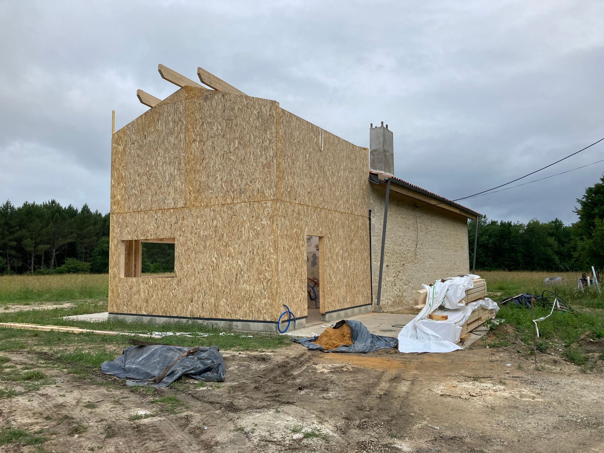Maison en construction avec murs en panneaux de particules ; cheminée et maçonnerie existante visibles.