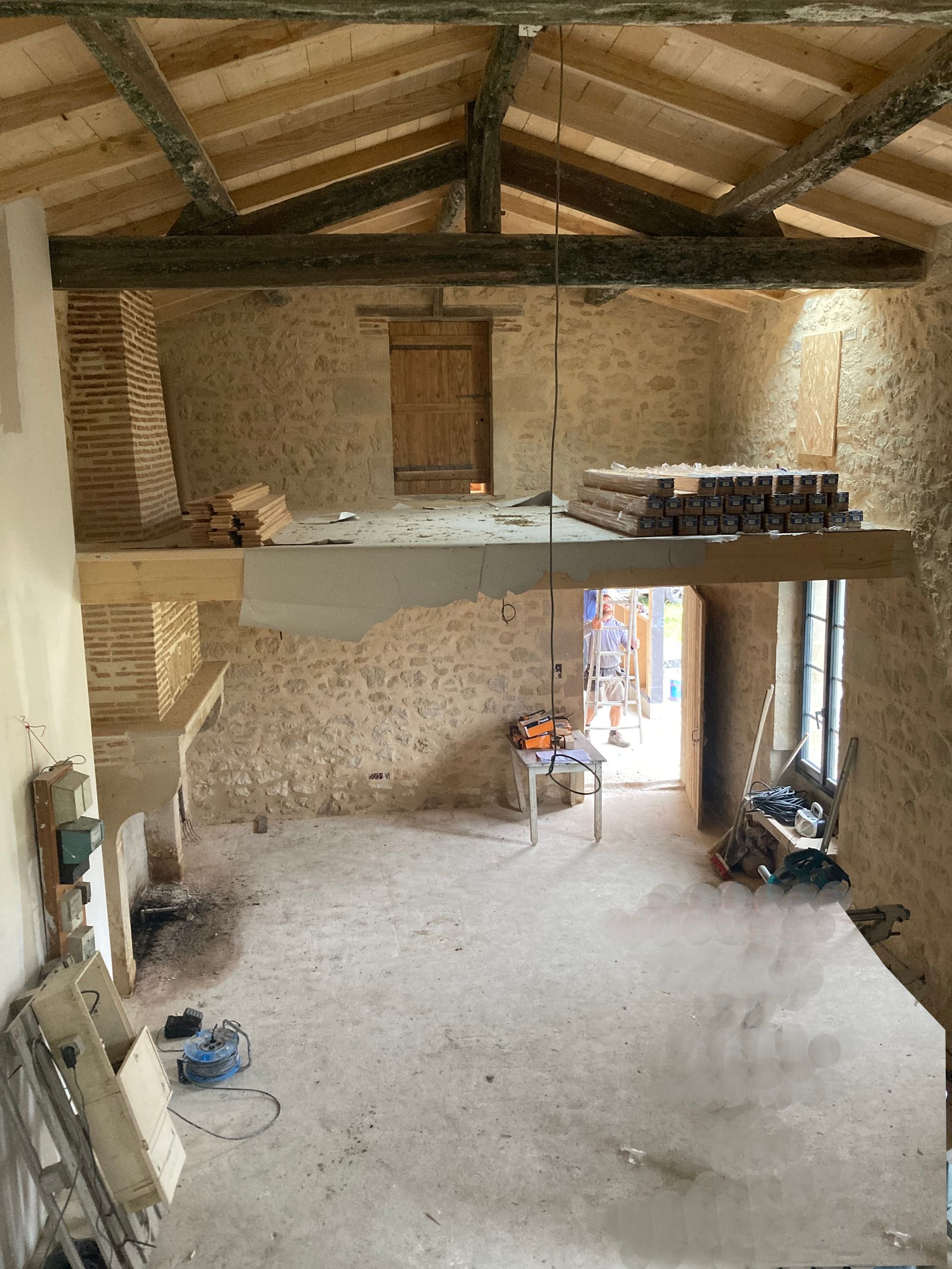 Rénovation intérieure : poutres apparentes, murs en pierre, deuxième étage partiel avec boiseries, outils, porte ouverte.