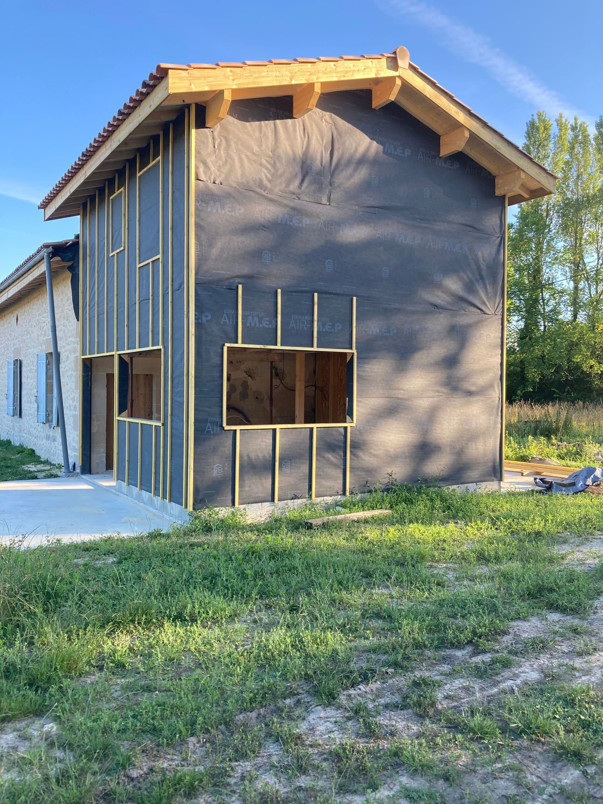 Extérieur du bâtiment en construction ; pare-vapeur noir, charpente en bois, fenêtres non finies et terrasse en béton.