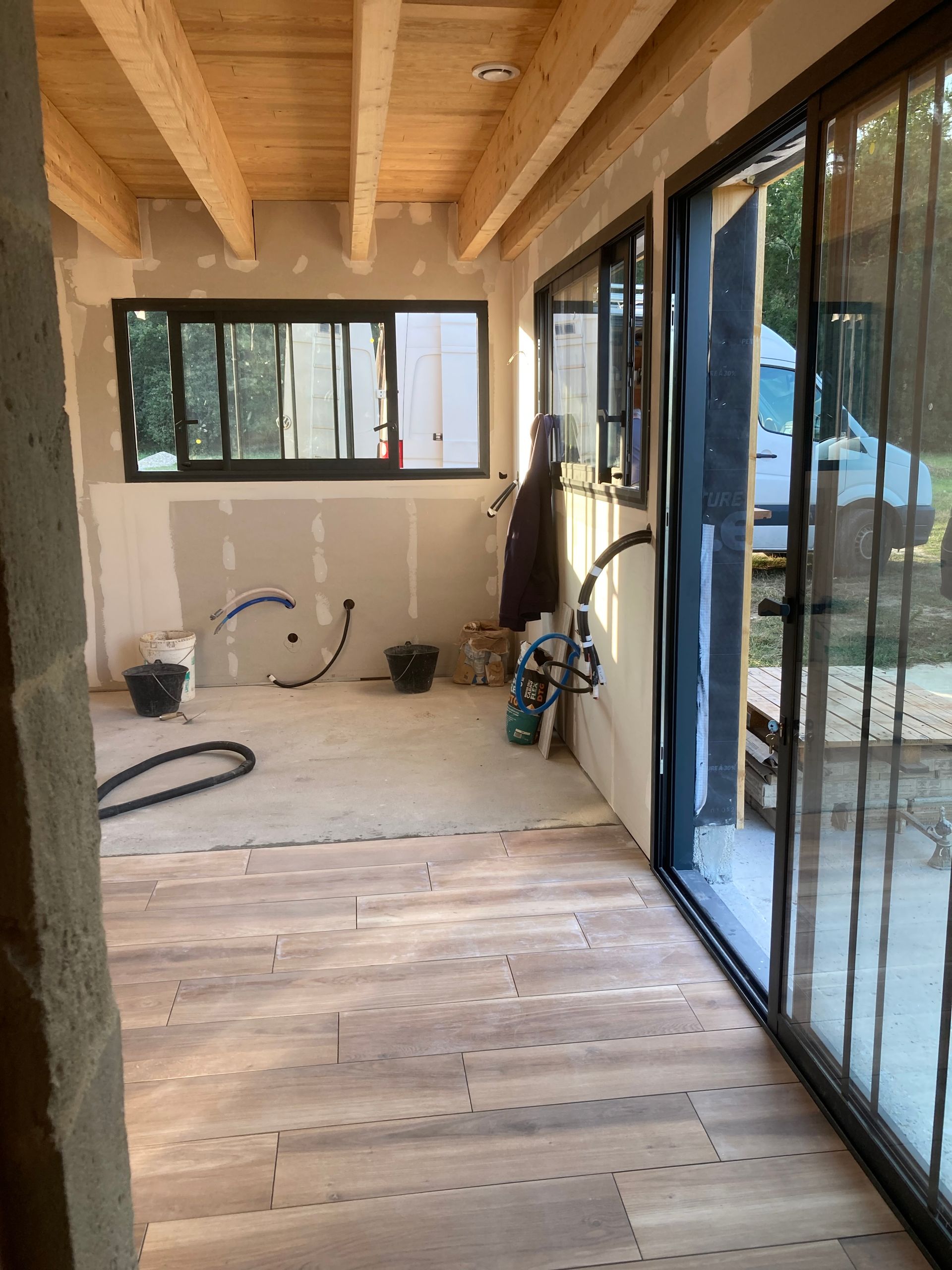 Pièce intérieure en cours d'aménagement avec poutres apparentes, cloisons sèches et grandes fenêtres. Le revêtement de sol est partiellement posé.