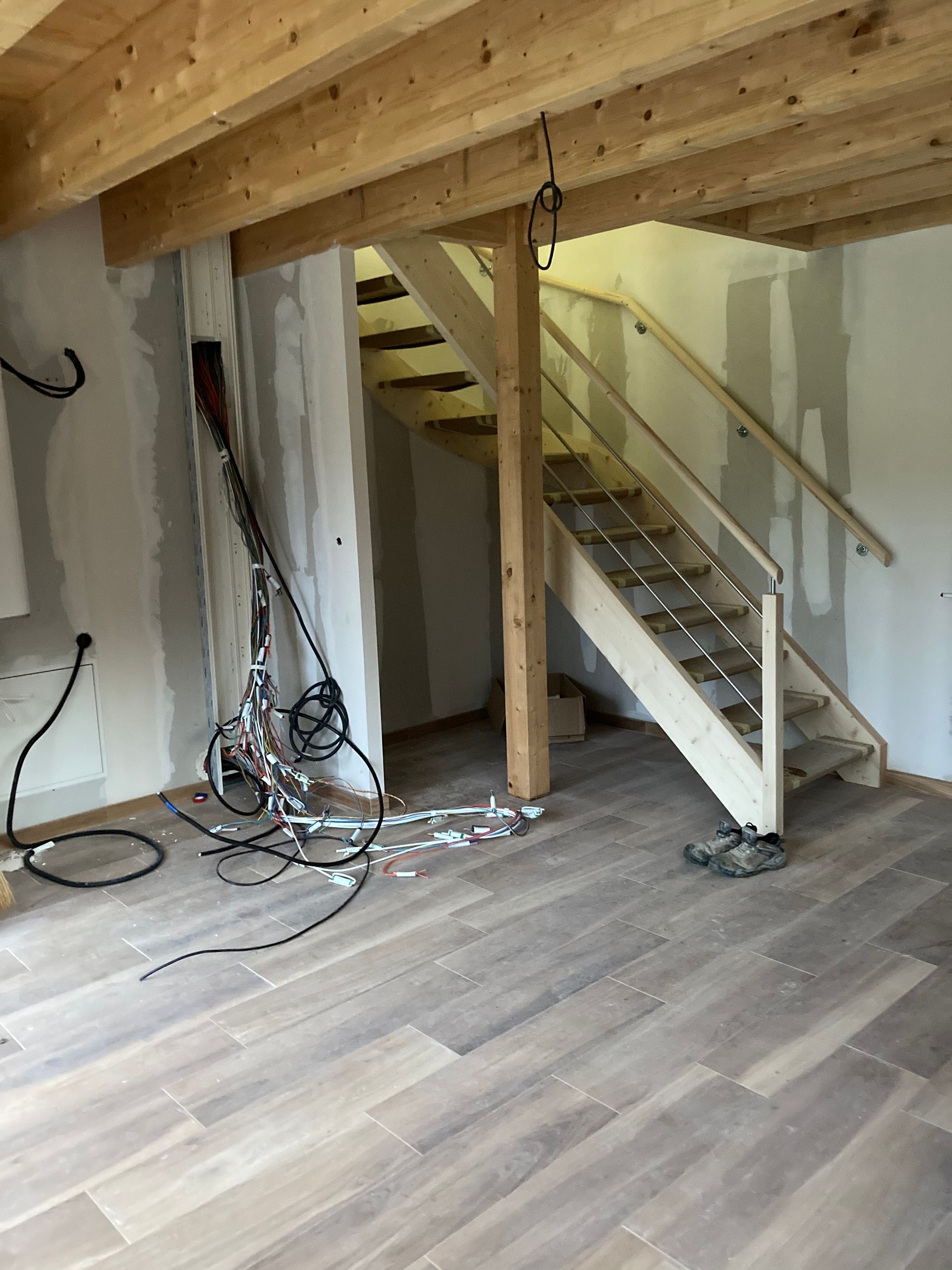 Vue intérieure : pièce inachevée avec escalier en bois, poutres apparentes et sol ; câblage électrique visible.