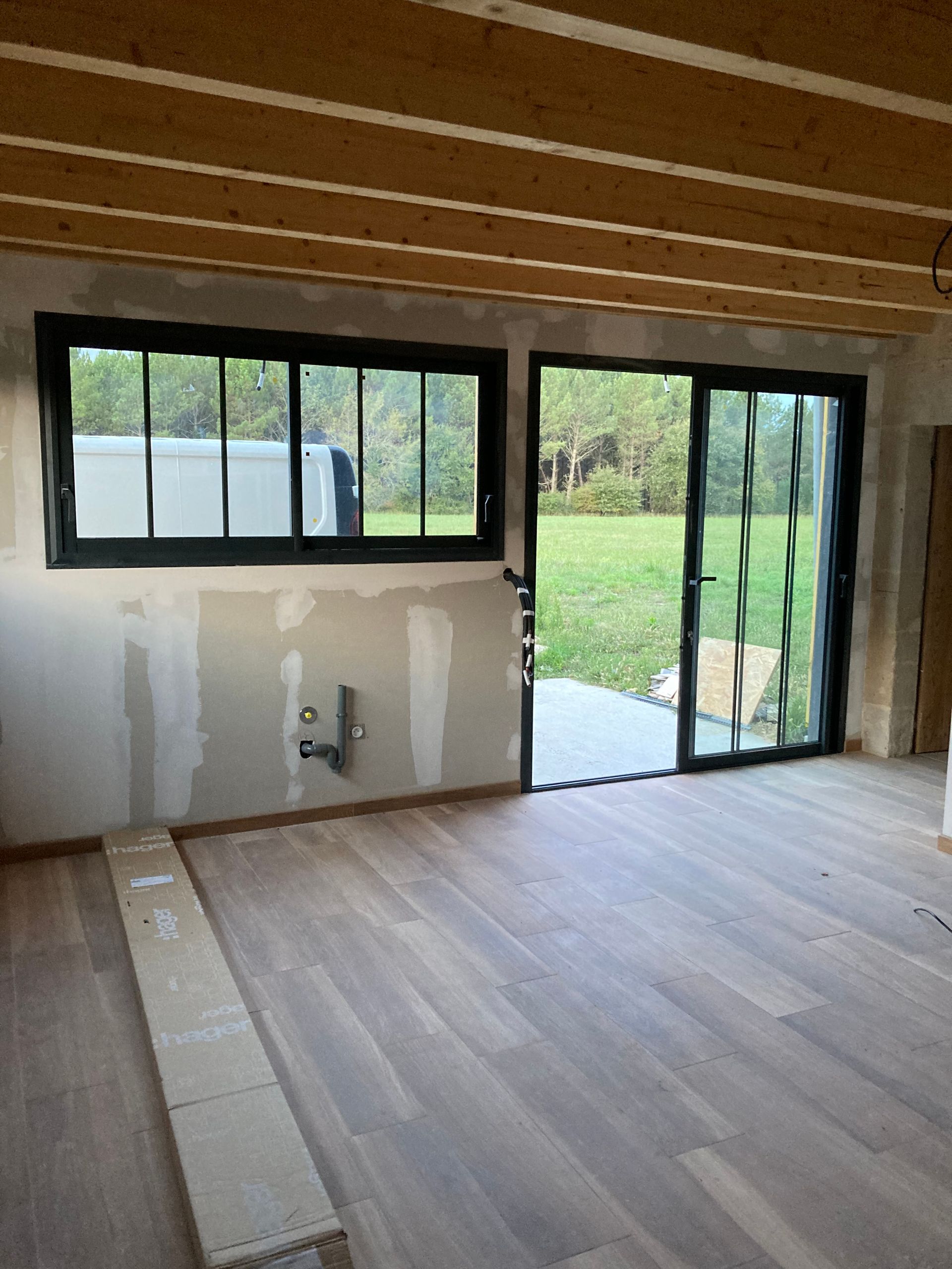 Intérieur d'une pièce avec fenêtres et porte à cadre noir, parquet, murs bruts et plafond en bois.