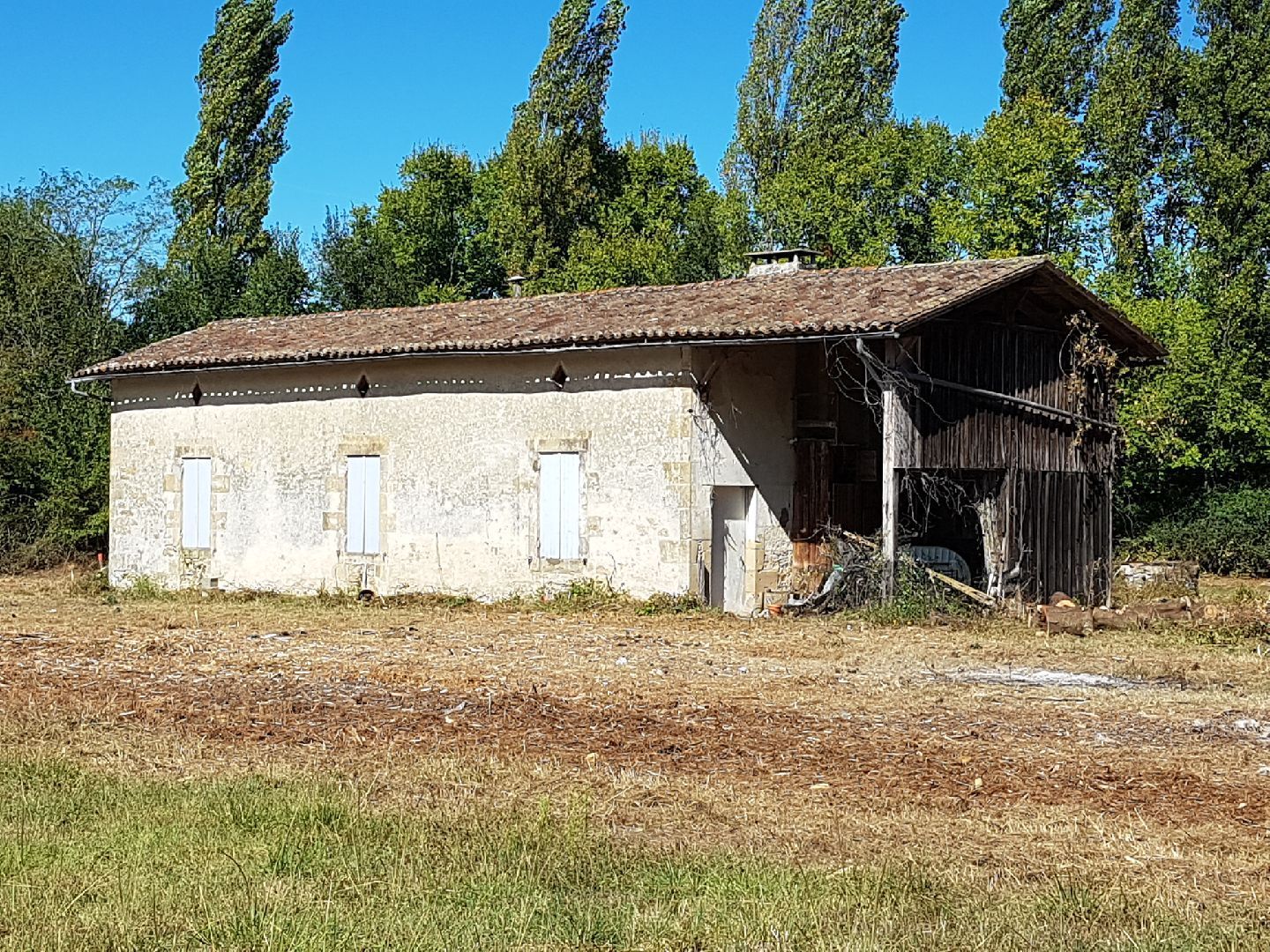 Un vieux bâtiment en pierre et en bois, usé par le temps, situé dans un champ, avec des volets fermés et des arbres en arrière-plan.