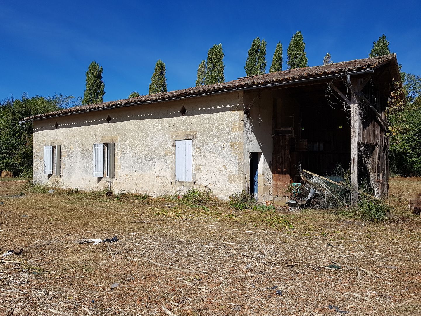 Bâtiment en pierre délabré, portes ouvertes, toit de bois usé par le temps et herbe sèche sous un ciel bleu.