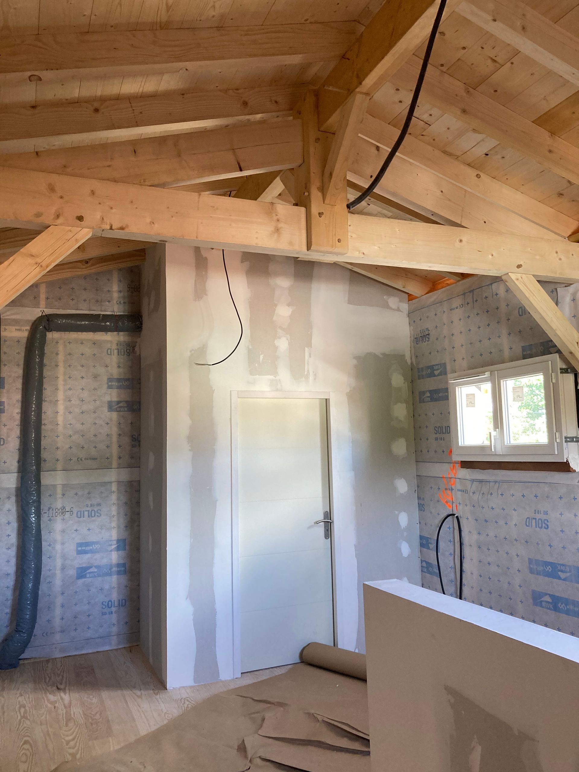 Pièce intérieure en construction, avec poutres en bois, isolation et une porte encadrée de plaques de plâtre.
