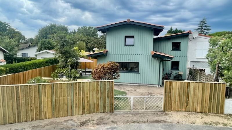 Maison verte avec clôture et portail en bois. Ciel nuageux, arbres en arrière-plan.