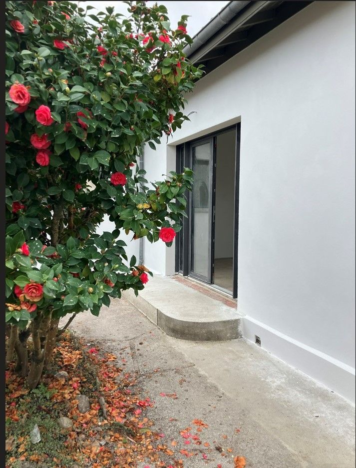 Un arbuste à fleurs rouges jouxte un bâtiment blanc doté de portes coulissantes en verre et d'une rampe en béton.