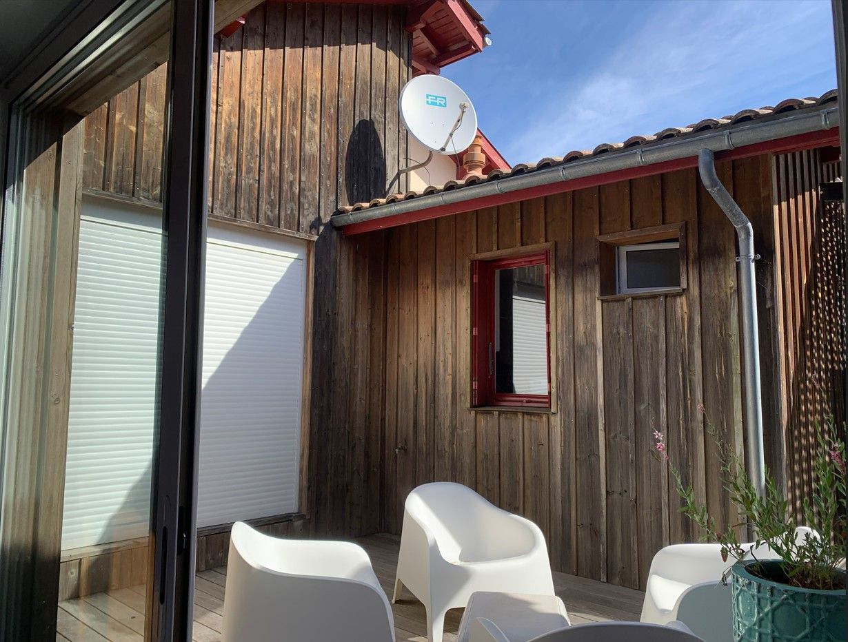 Terrasse avec murs en bois, chaises blanches et antenne parabolique. Journée ensoleillée.