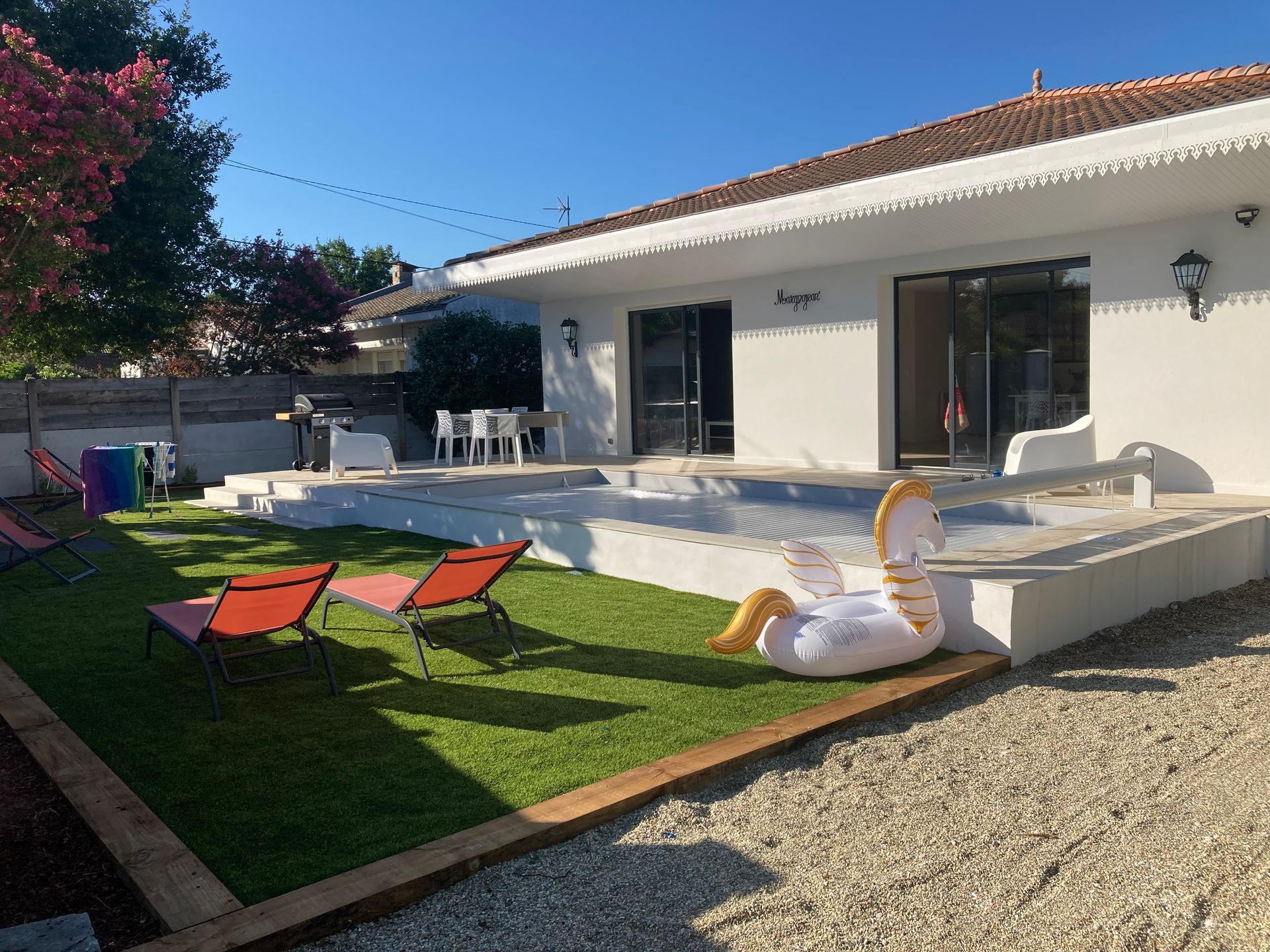 Jardin avec piscine, chaises longues, licorne gonflable et maison blanche avec portes coulissantes en verre.