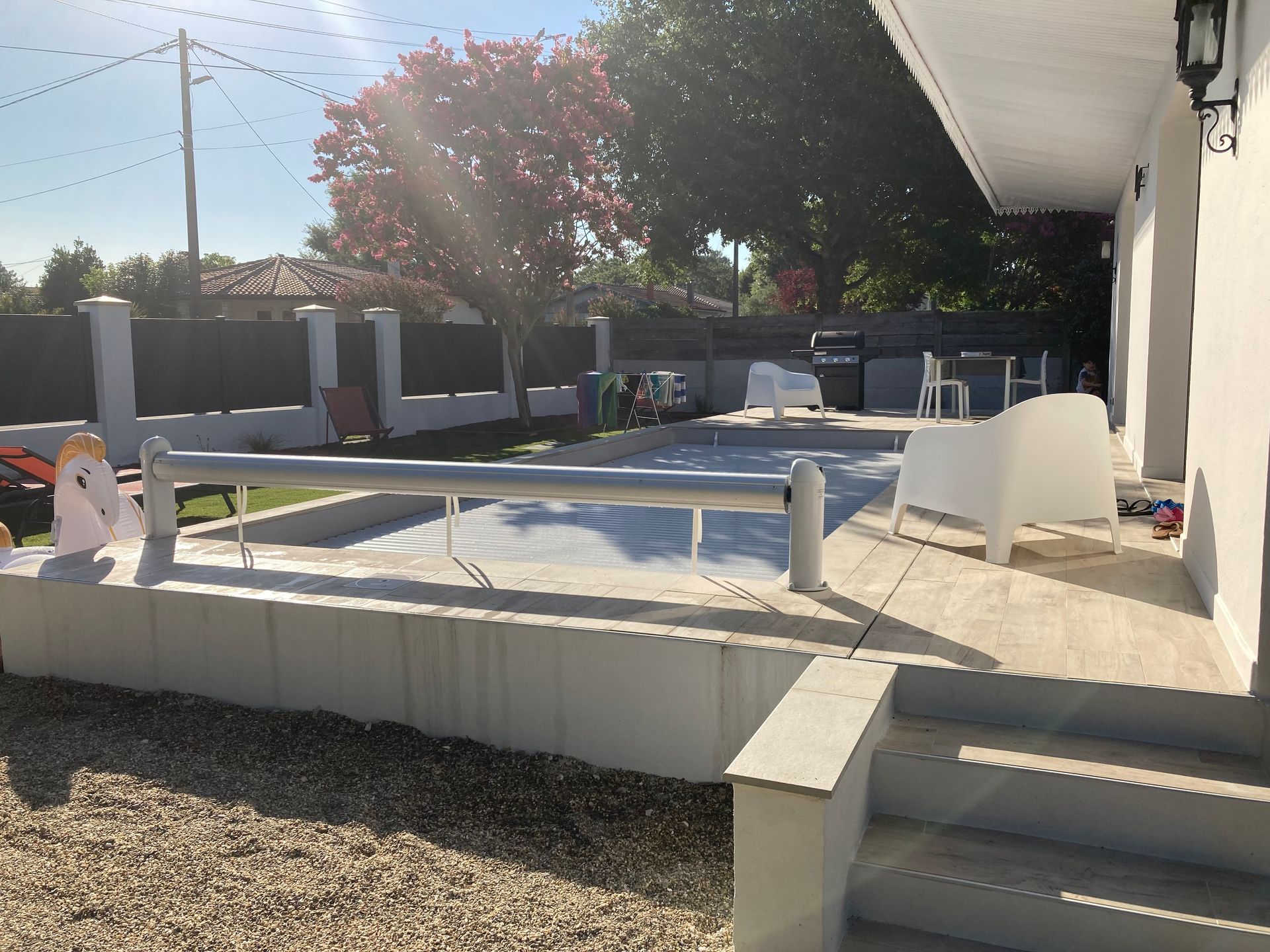 Un jardin avec piscine. Du mobilier de jardin blanc est disposé près de la piscine. Un mur gris entoure la piscine.