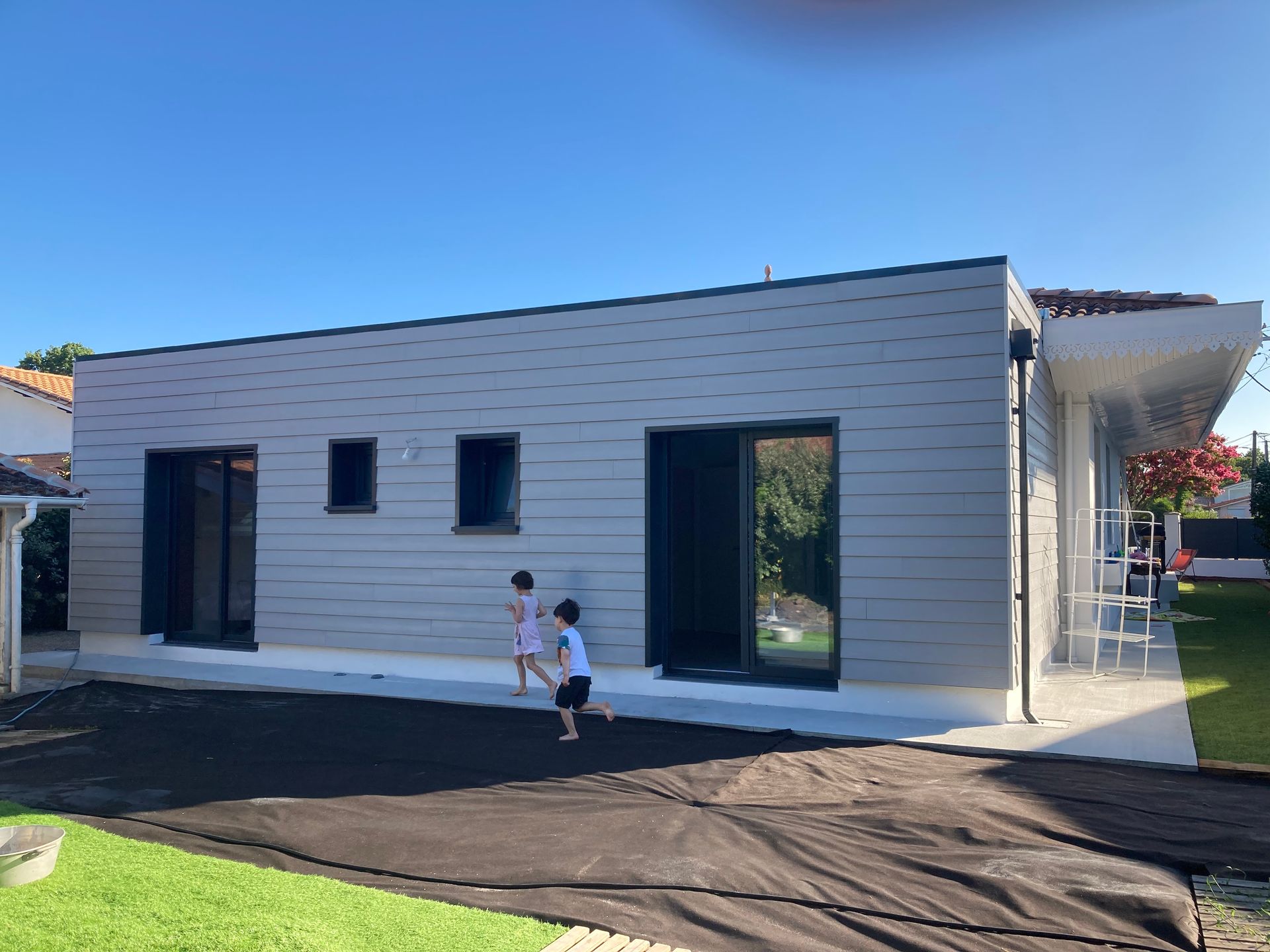 Des enfants jouent près d'une maison grise moderne aux portes et fenêtres à cadre noir, par une journée ensoleillée.