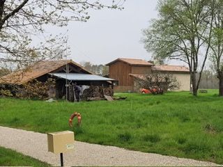 Des bâtiments se dressent au milieu d'un champ herbeux ; on aperçoit des tas de bois, un chemin de gravier et une voiture orange. Ciel nuageux.