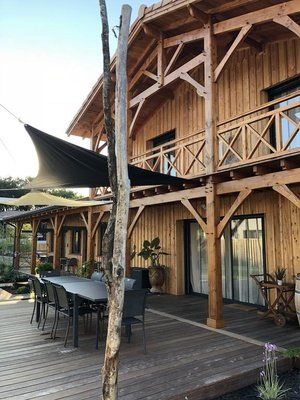 Maison en bois avec terrasse, patio ombragé avec table et chaises. Voiles d'ombrage noires, arbres et plantes.