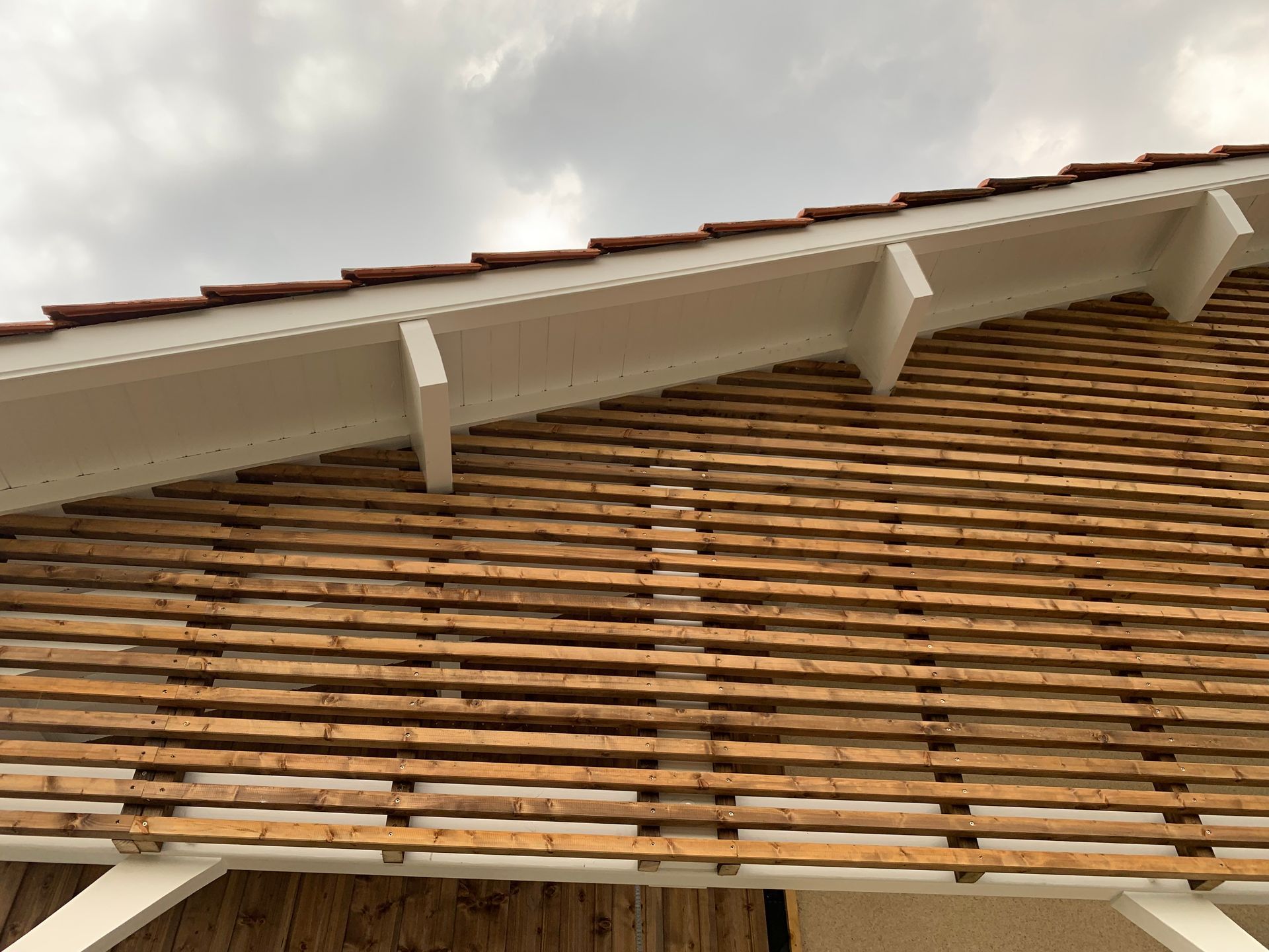 Lattes de bois sur une charpente de toit, avec des finitions blanches et un ciel nuageux au-dessus.