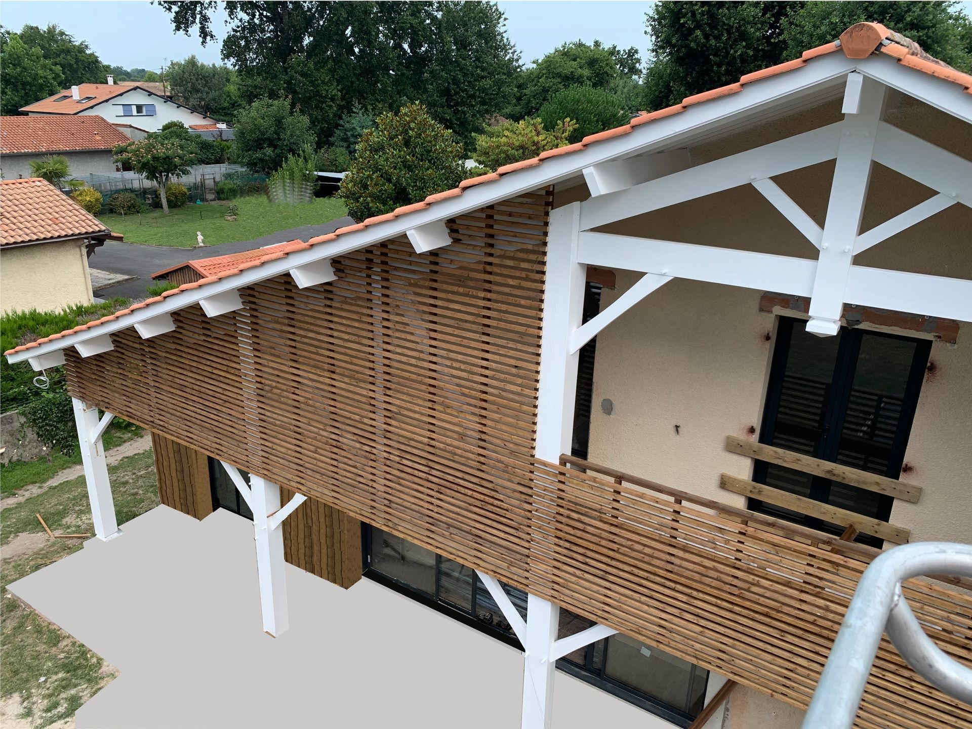 Maison à deux étages avec bardage en treillis de bois et boiseries blanches. Toit de tuiles rouges, balcon et arbres en arrière-plan.