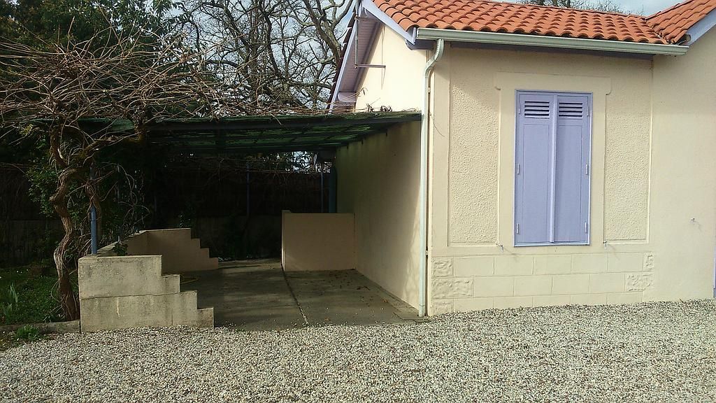 Maison beige au toit de tuiles rouges, abri voiture et volets bleus. Cour en gravier gris, marches et arbre sans feuilles.