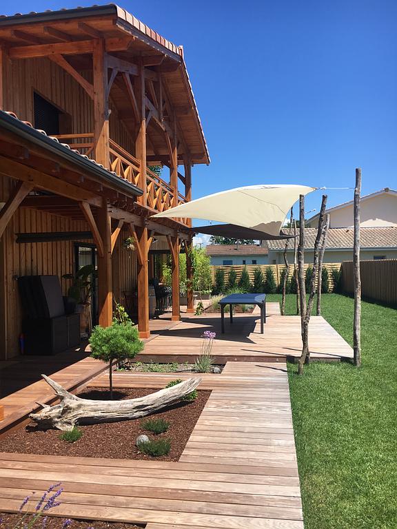 Terrasse en bois et maison avec un voile d'ombrage au-dessus d'une table, journée ensoleillée.