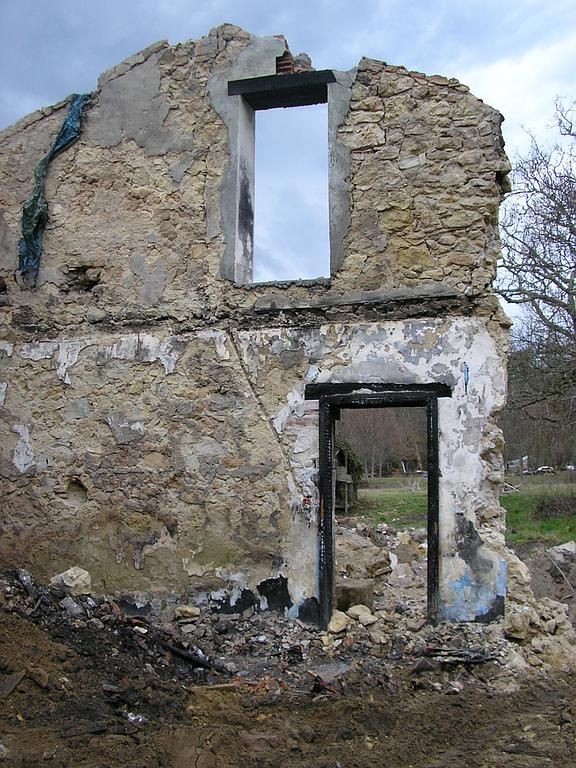 Façade en pierre délabrée, avec encadrements de fenêtres et de portes vides ; débris à la base.