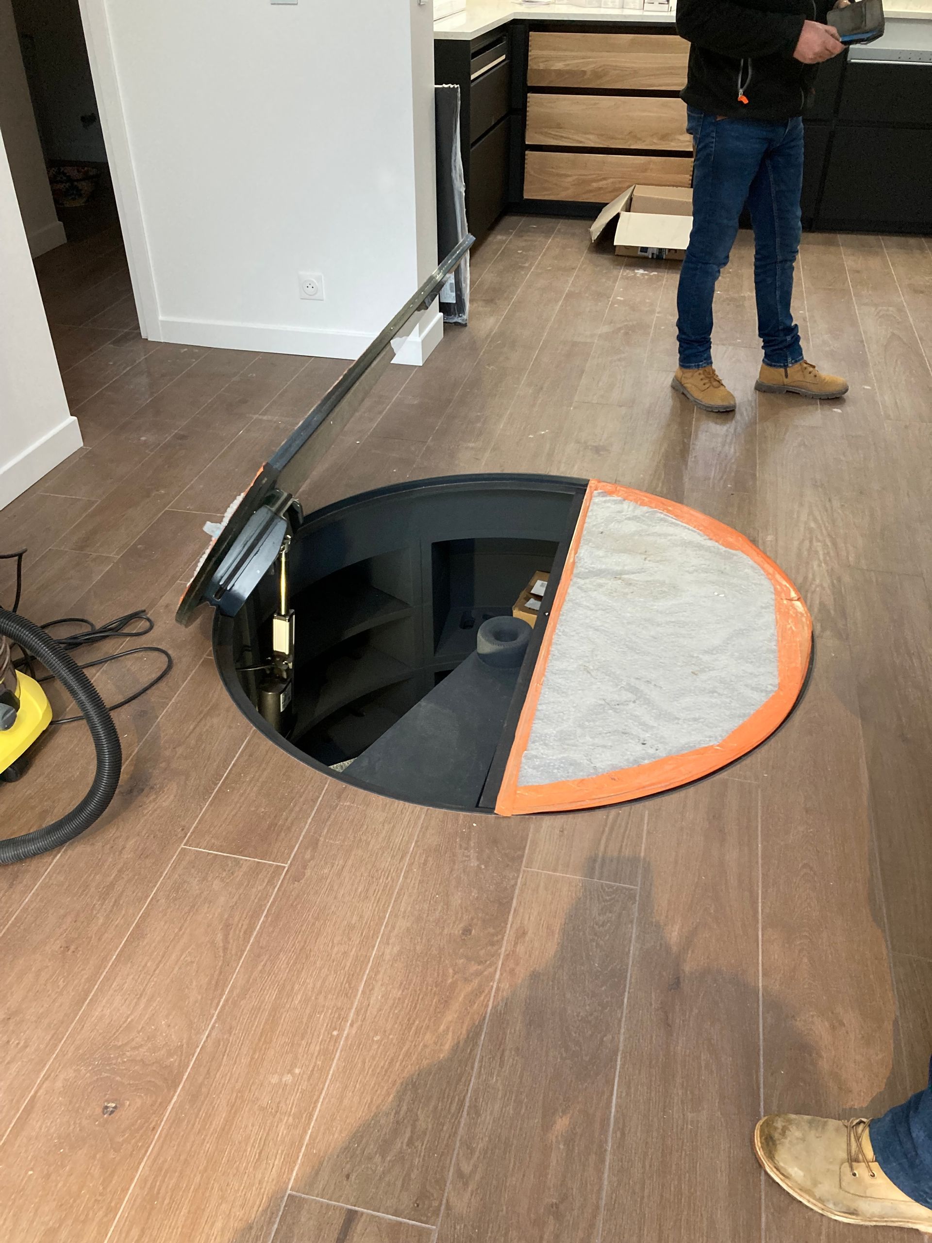 Trappe circulaire ouverte au sol donnant accès à un escalier menant à un niveau inférieur. Une personne se tient à proximité. Sol en bois.