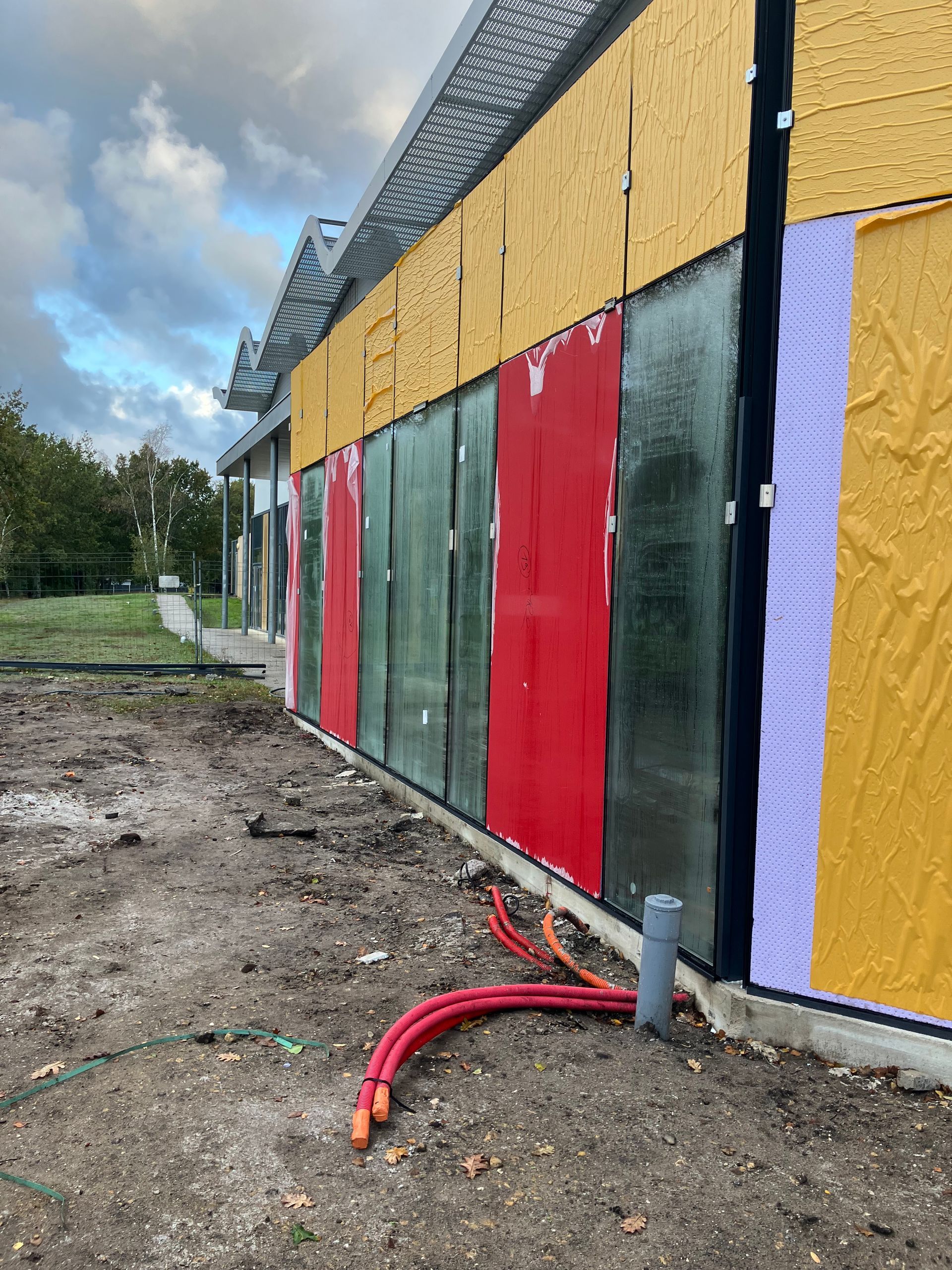 Bâtiment en construction avec contreplaqué apparent, panneaux rouges et isolation. Des tuyaux rouges sortent du bâtiment.