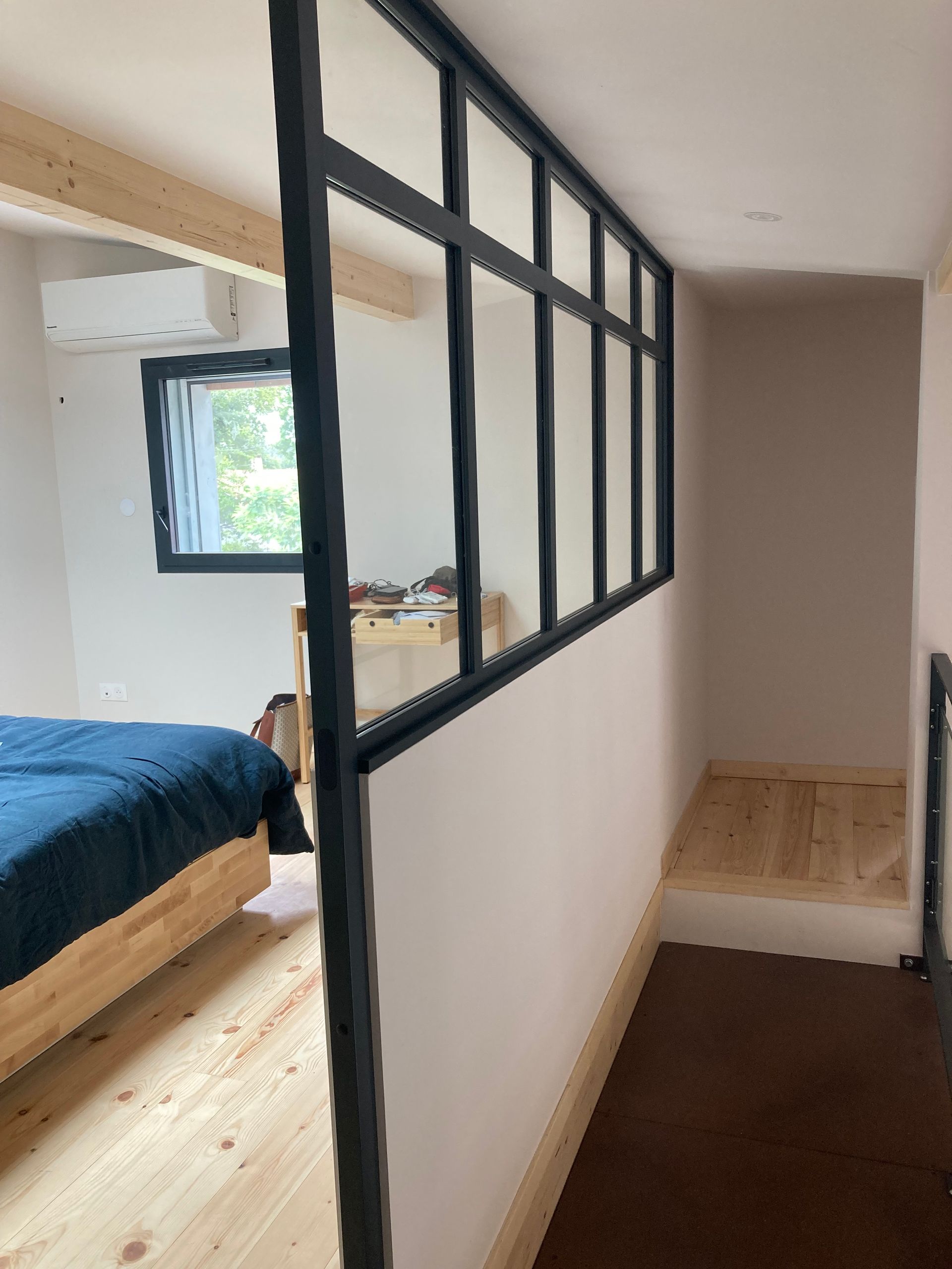 Cloison vitrée à cadre noir séparant une chambre. Lit en bois, fenêtre et murs aux tons neutres.
