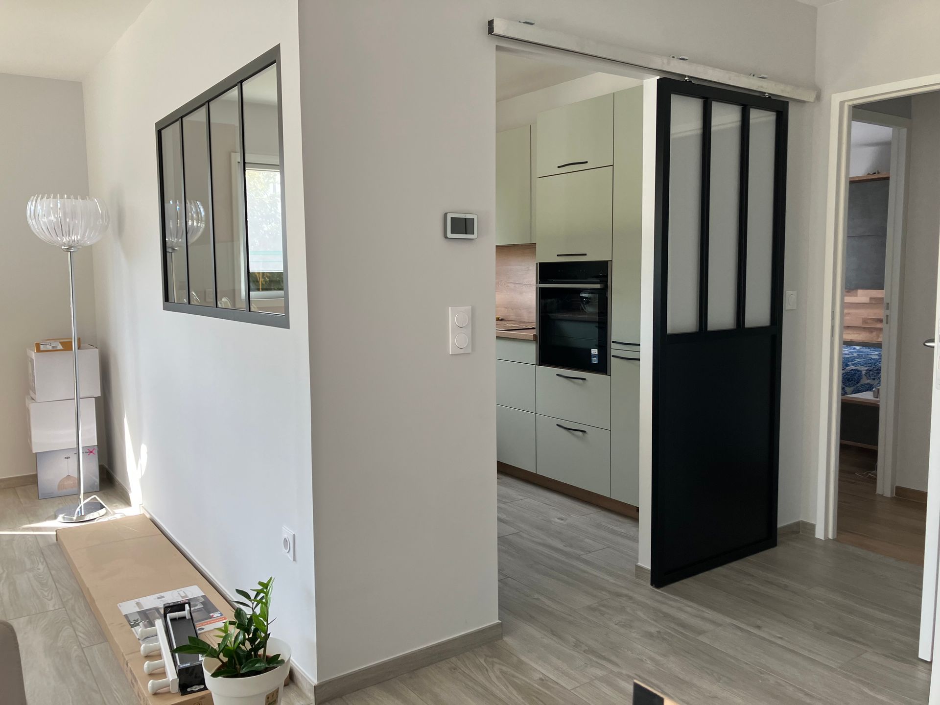 Une porte coulissante en métal noir donne sur la cuisine ; murs blancs, sol gris, fenêtre à gauche.