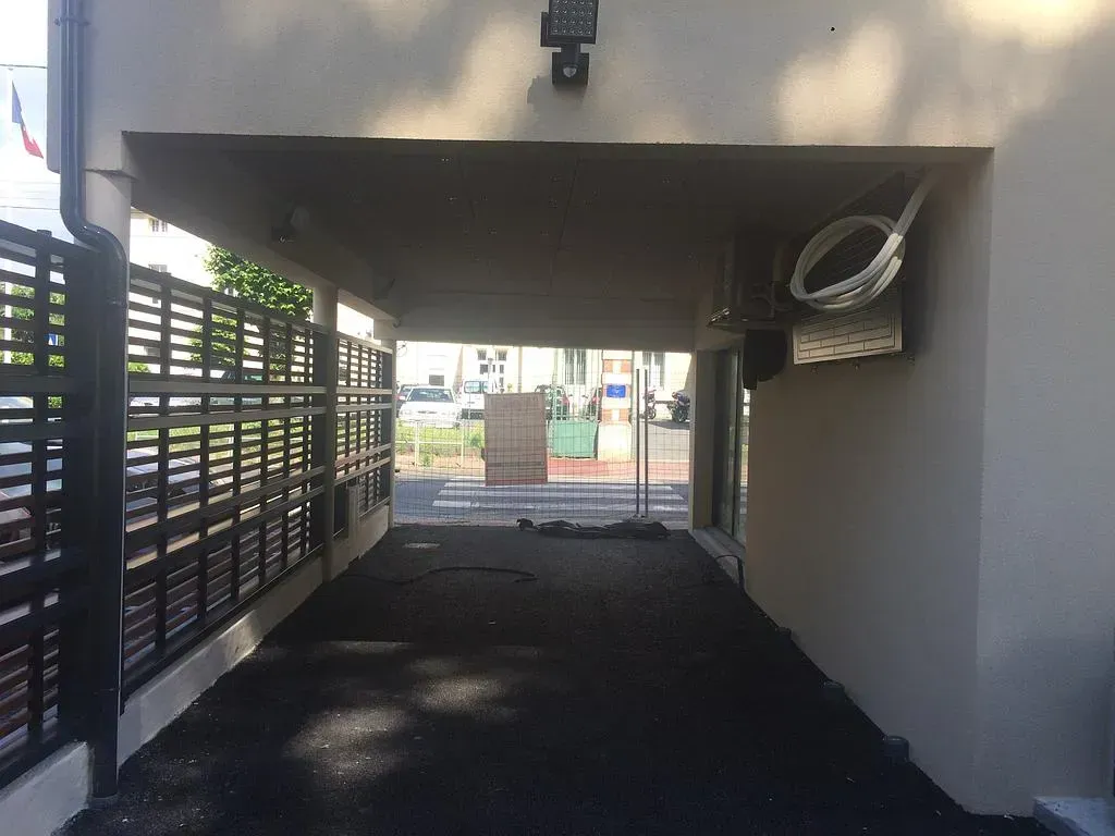 Allée carrossable sous un auvent. Asphalte noir. Clôture à gauche. Lumière en haut de l'auvent.