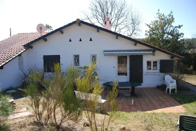 Maisonnette blanche avec un toit de tuiles, des volets noirs et une petite terrasse avec mobilier de jardin.