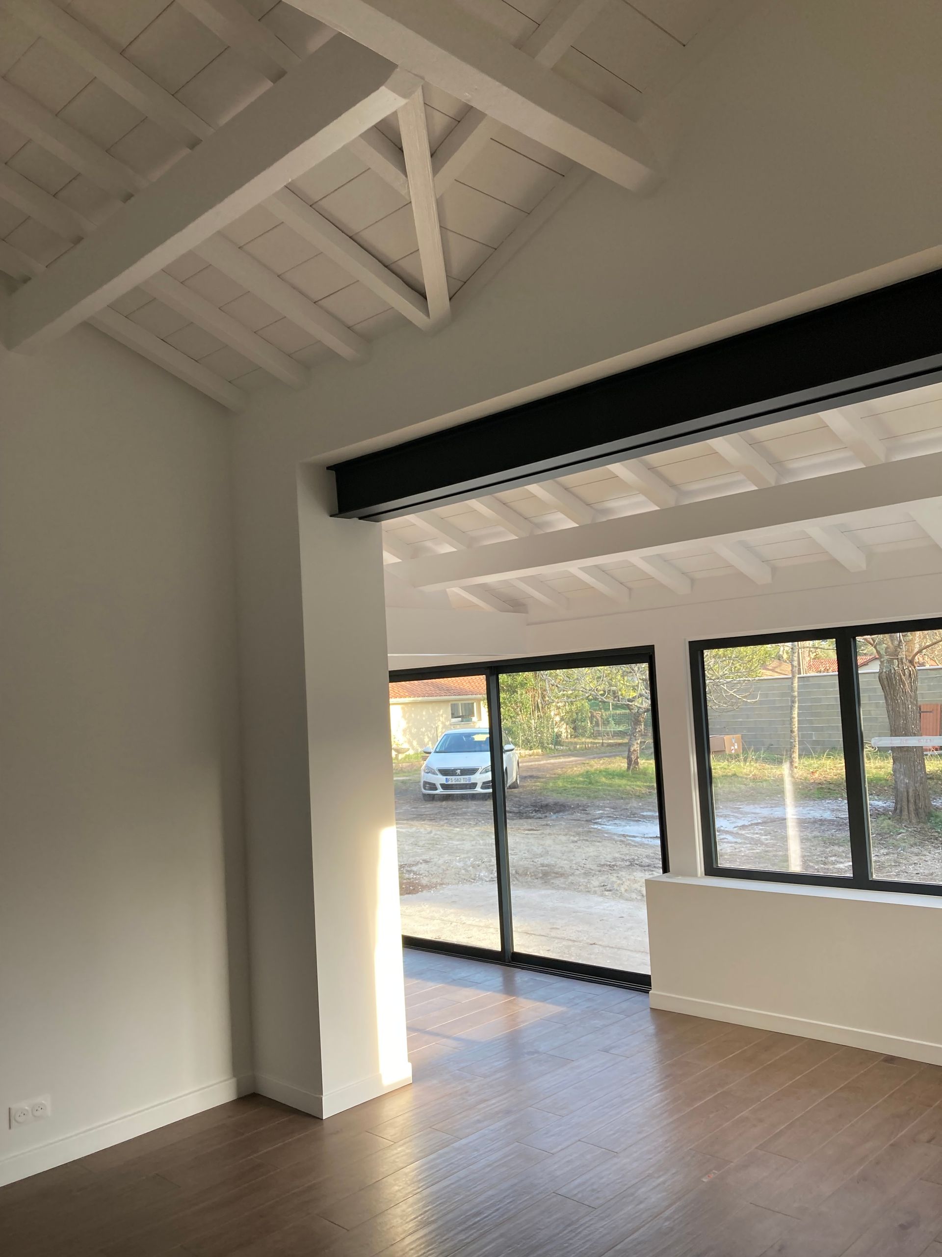 Vue intérieure d'une pièce aux murs blancs, poutres apparentes et grandes fenêtres à cadre noir ; une voiture est visible à l'extérieur.