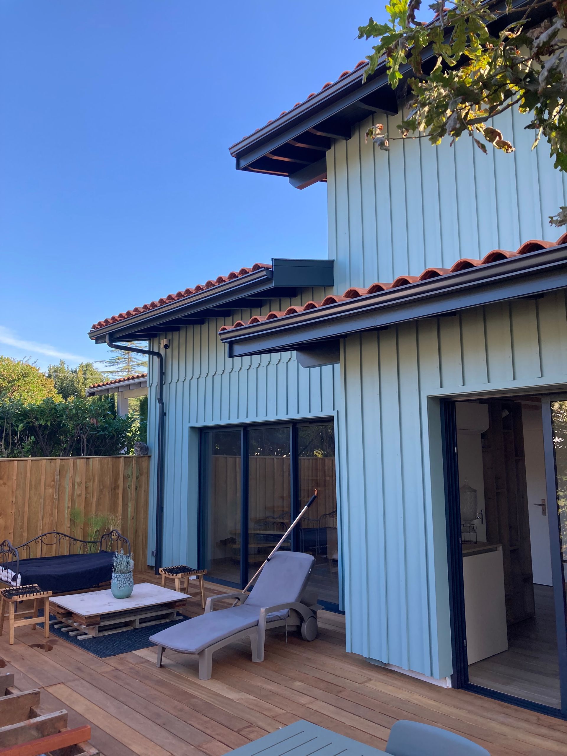 Maison aux murs bleus avec terrasse en bois et mobilier de jardin ; journée ensoleillée.