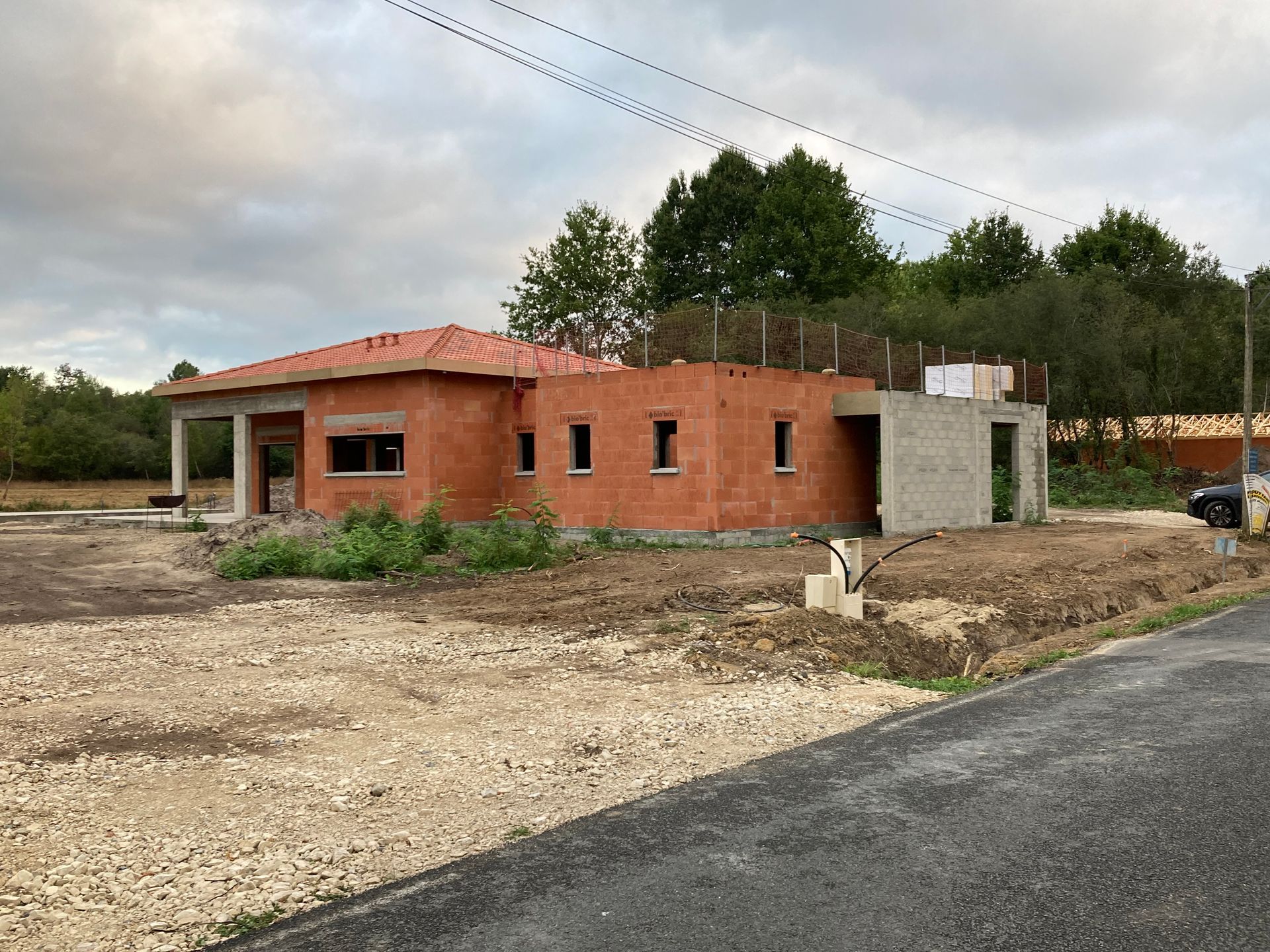 Une maison en briques partiellement construite, avec un toit de tuiles rouges, sur un terrain gravillonné près d'arbres et d'une route.