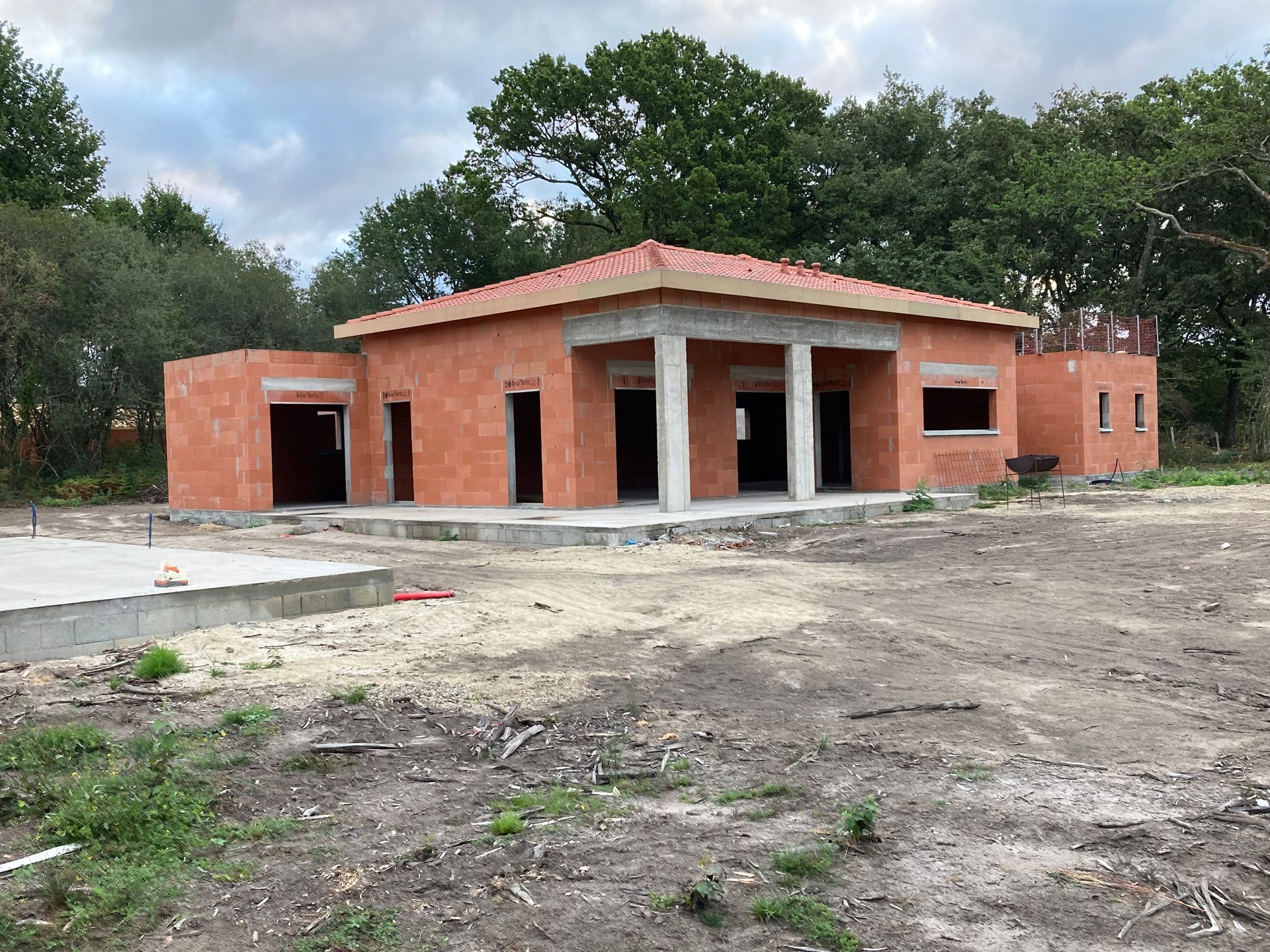 Bâtiment en construction, avec façade en briques rouge-brun, colonnes blanches et toit de tuiles orange, entouré de terre et d'arbres.