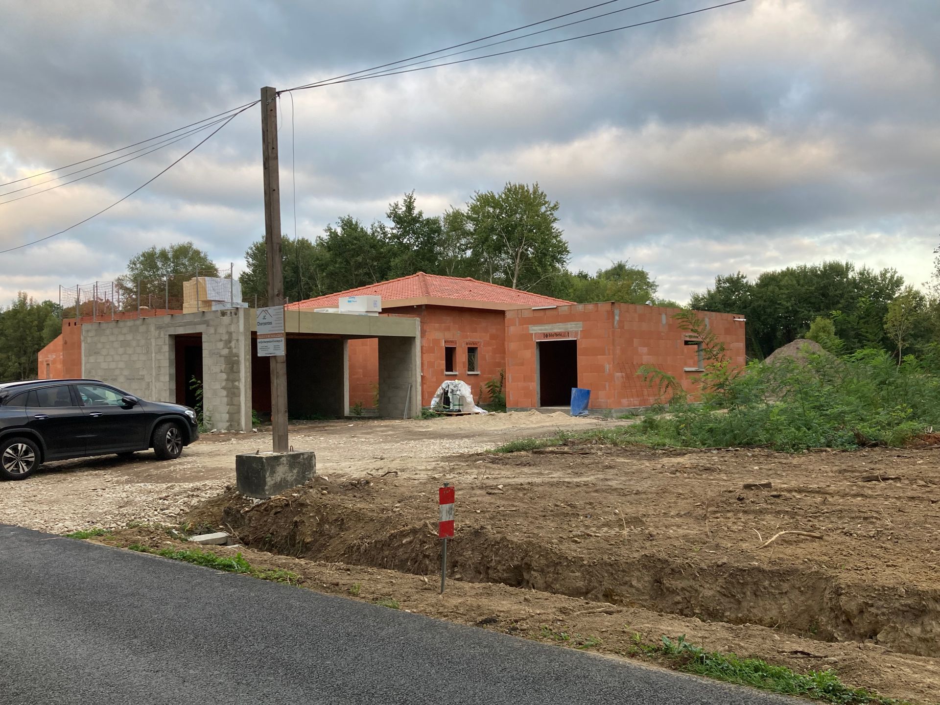 Une maison en construction, une voiture garée devant. Le ciel est nuageux et le terrain alentour est en terre.