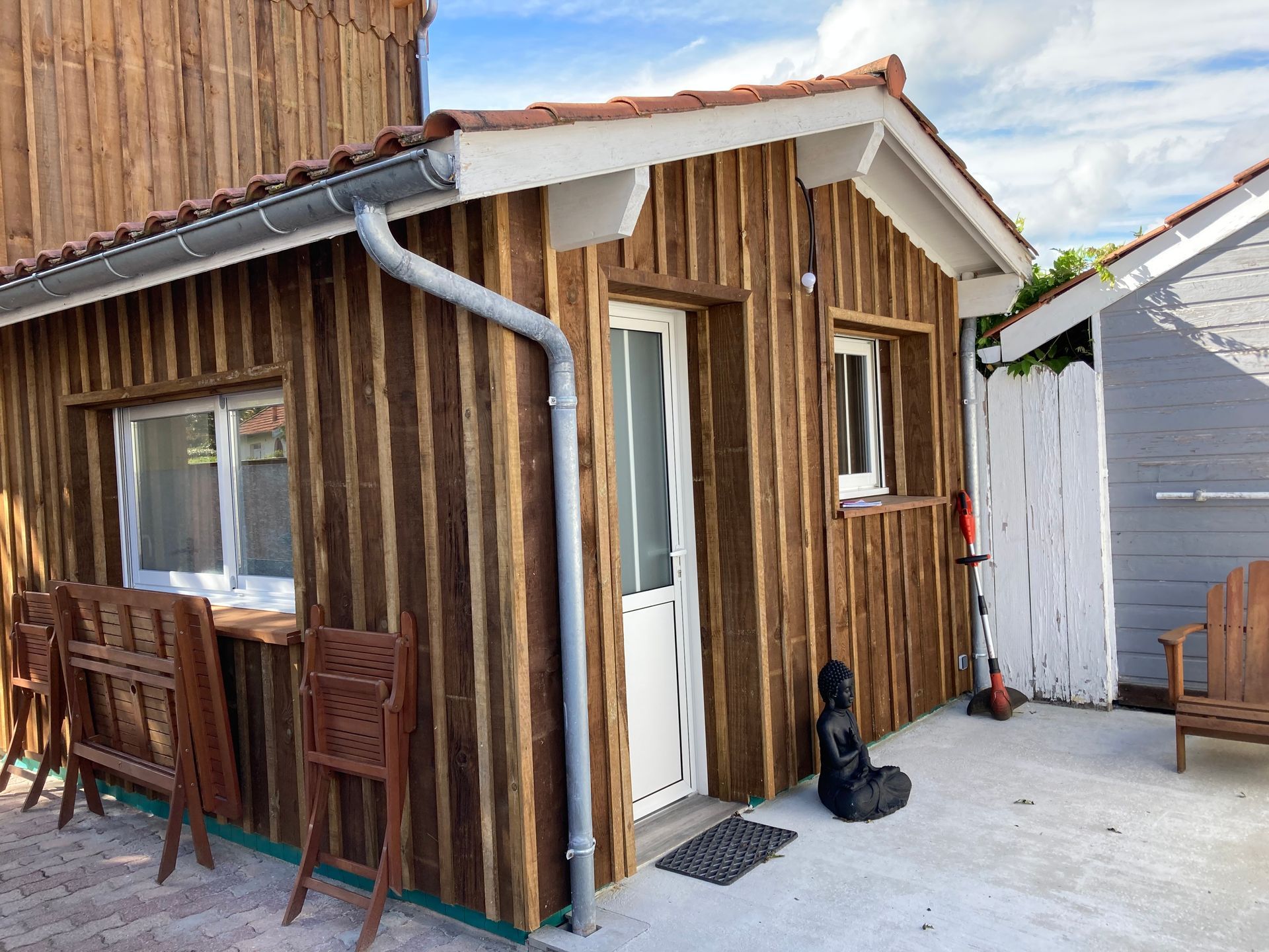 Cabane en bois avec porte et fenêtre blanches, gouttière grise, petit patio et statue de Bouddha.