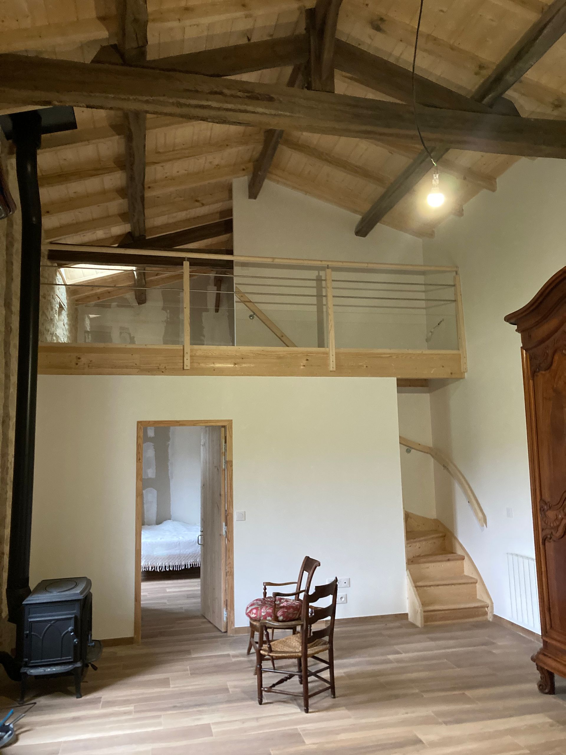 Intérieur d'une pièce rustique avec mezzanine et poêle à bois. On y trouve des poutres apparentes, un escalier et une chaise en bois.