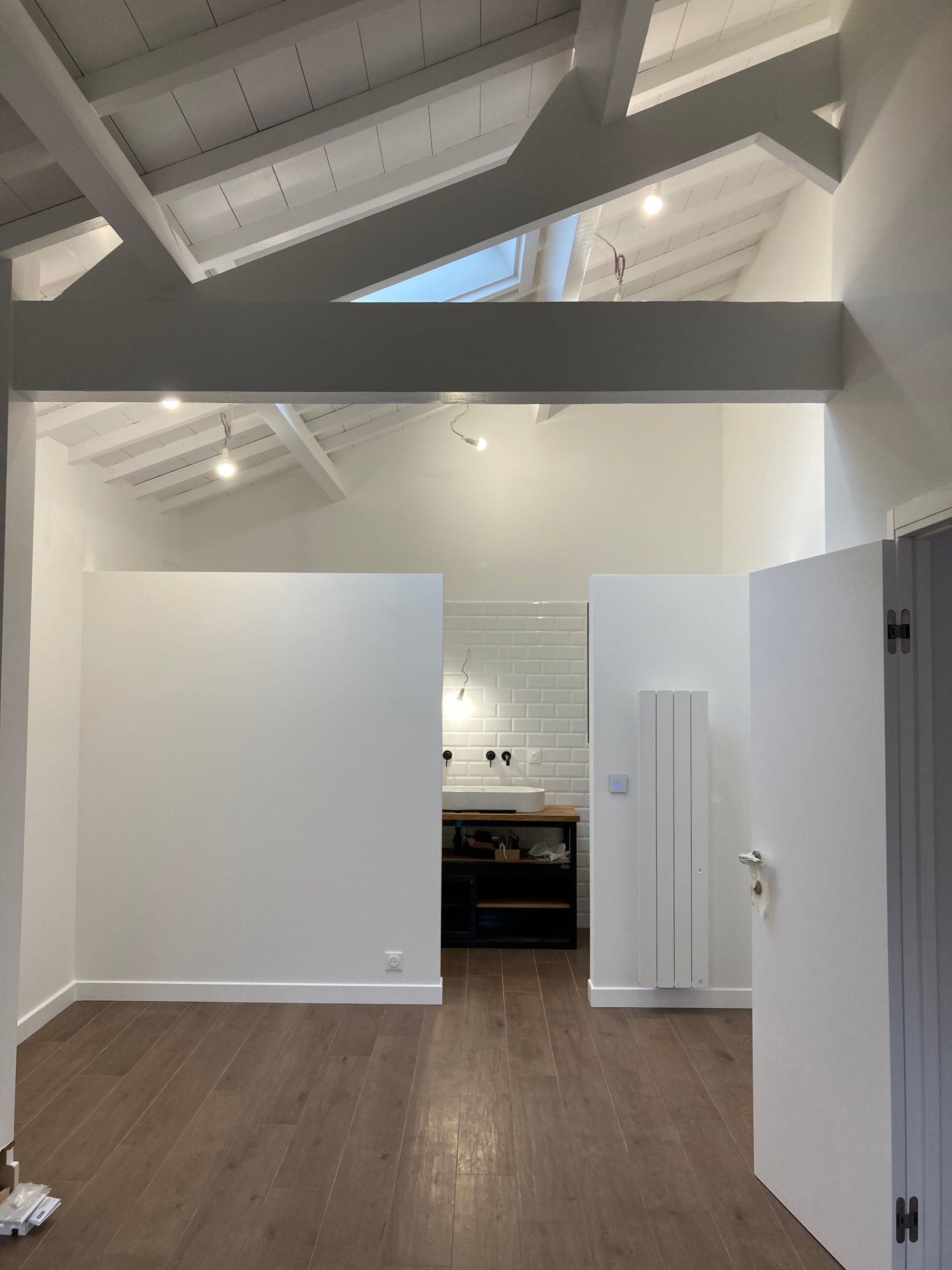 Vue intérieure d'un espace moderne avec parquet, murs blancs et poutres apparentes, menant à un coin cuisine.