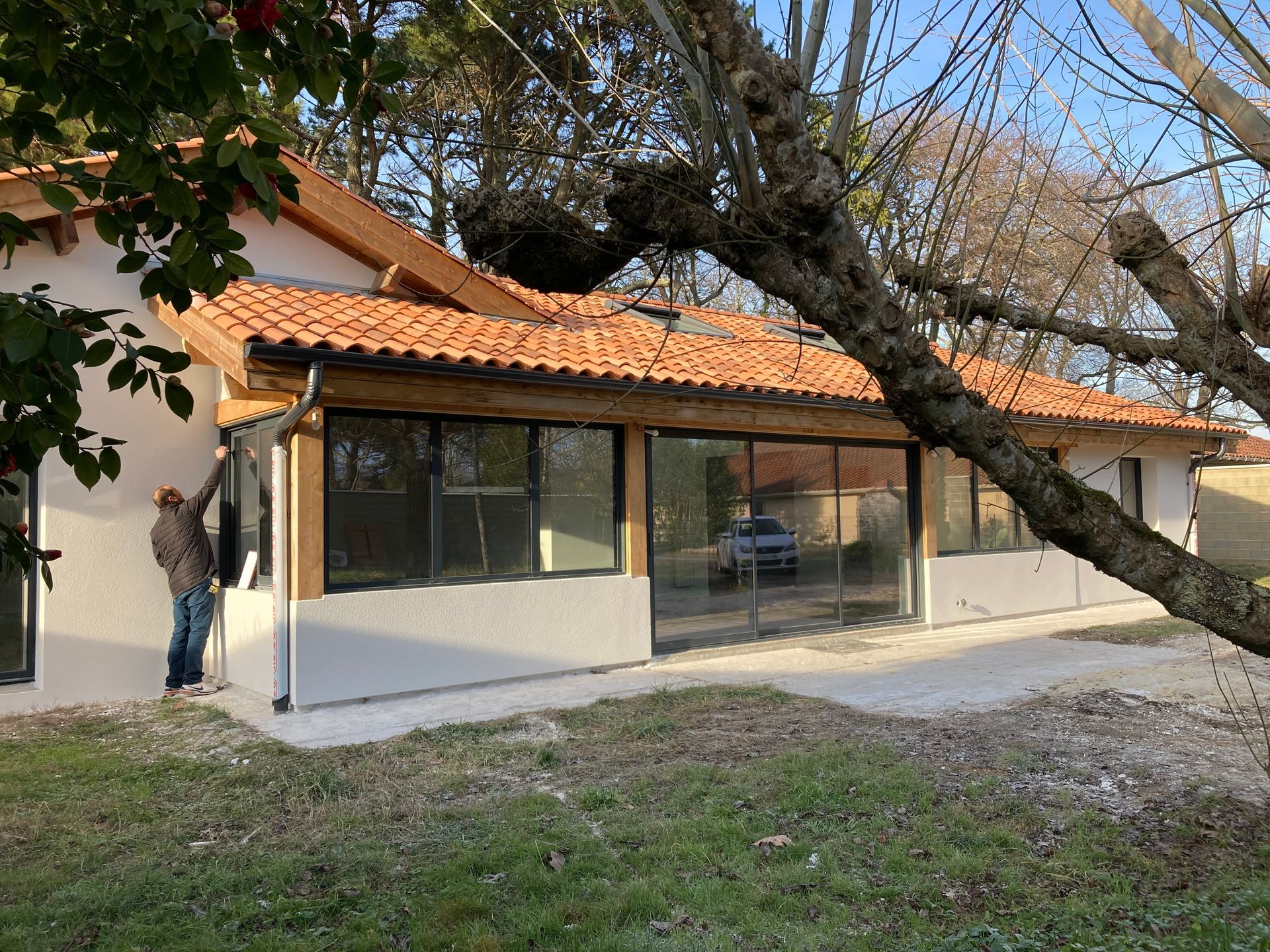 Un homme peint l'extérieur d'une maison au toit de tuiles rouges et aux grandes fenêtres, entourée d'arbres.