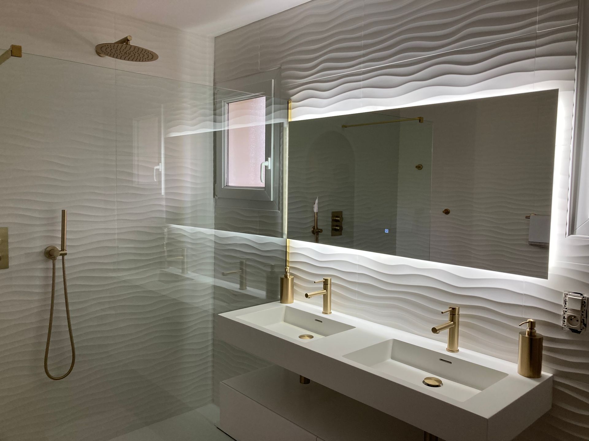 Salle de bains moderne avec murs blancs texturés, douche en verre, double vasque et robinetterie dorée.