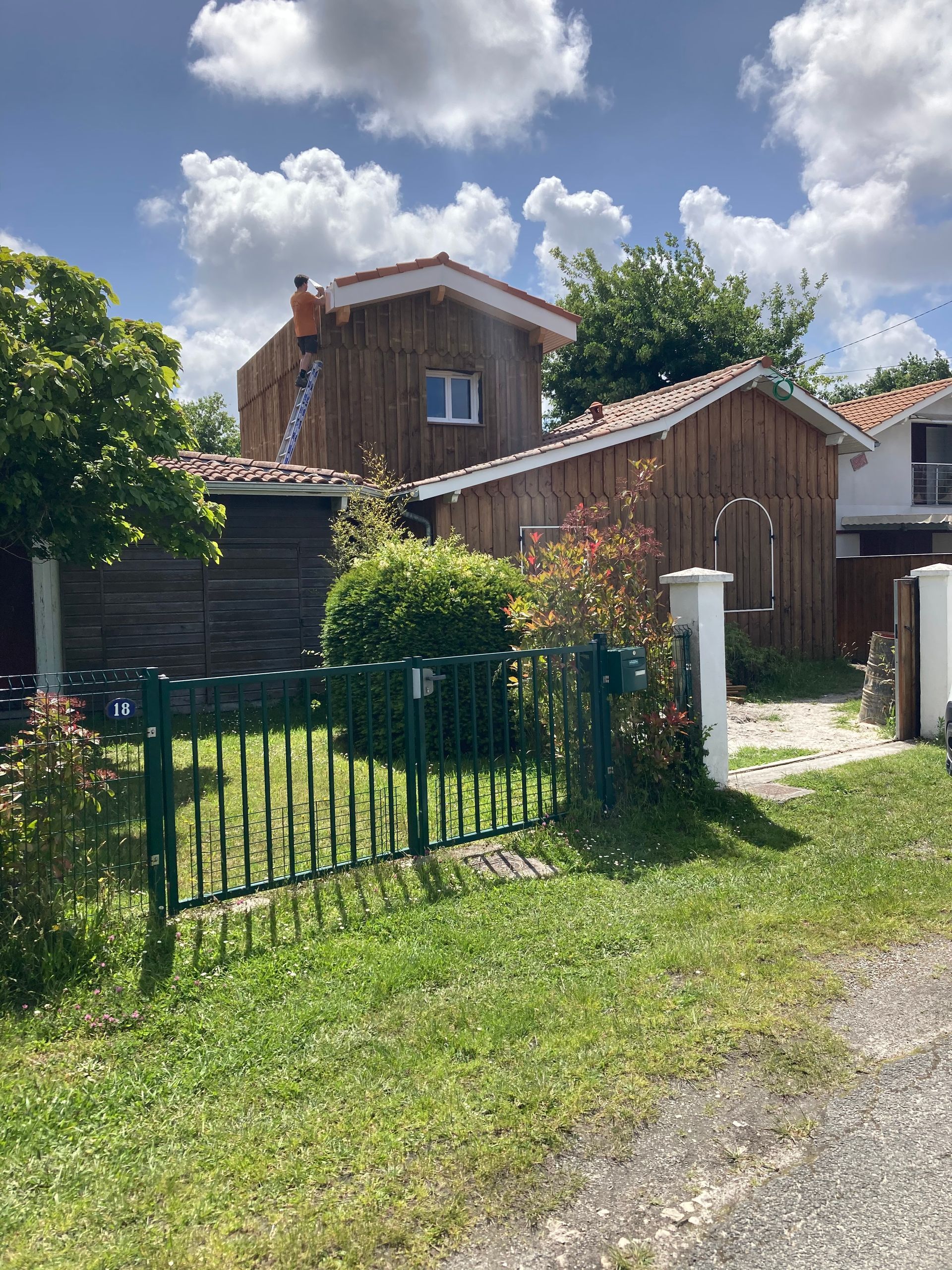 Maison en bois au toit rouge, à la clôture verte et au jardin verdoyant sous un ciel bleu partiellement nuageux.
