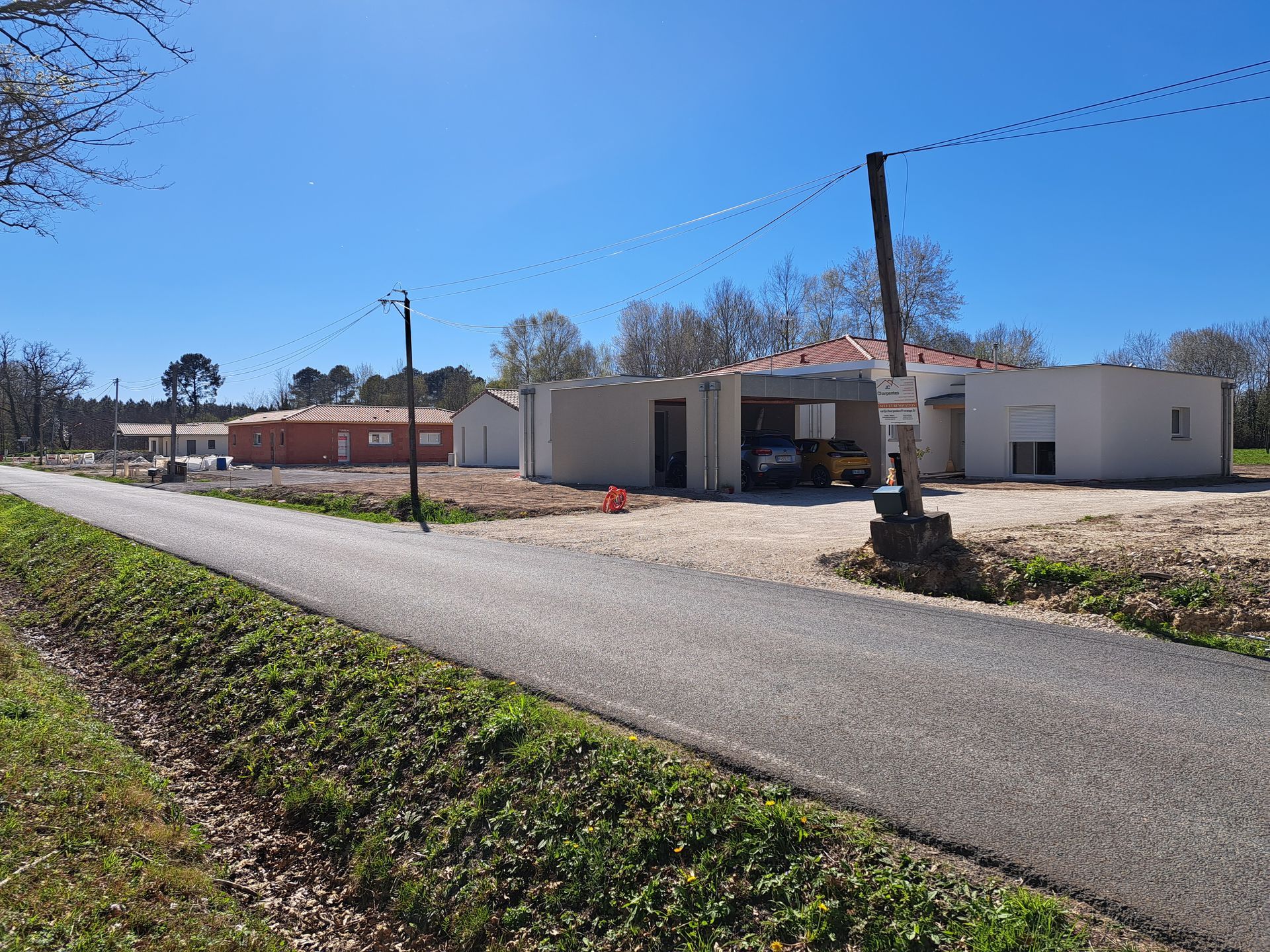 Chantier de construction avec plusieurs bâtiments, allée de gravier, poteau électrique, ciel bleu dégagé.