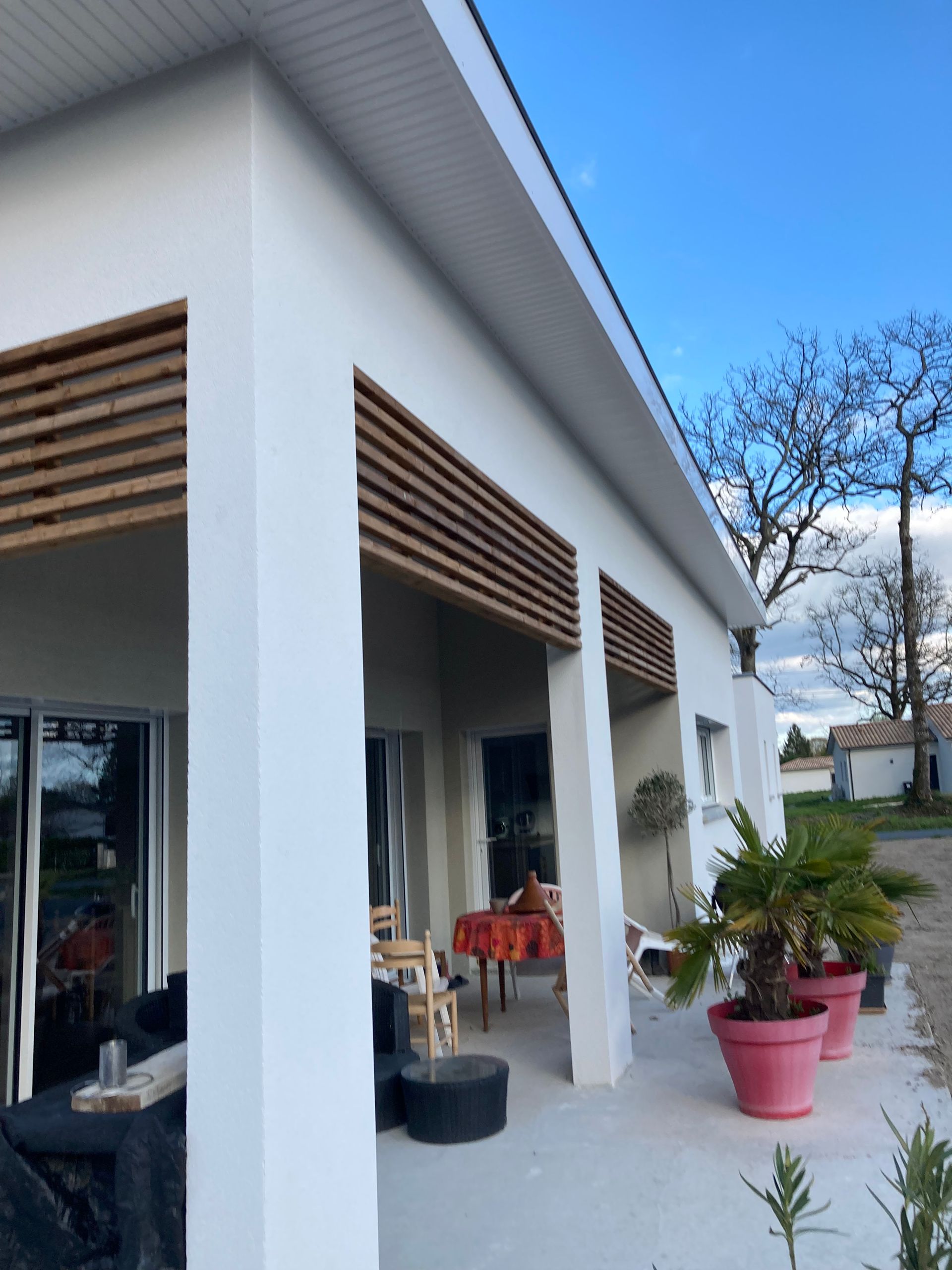 Maison blanche avec des lattes de bois, des plantes en pot et des sièges extérieurs sous un porche.