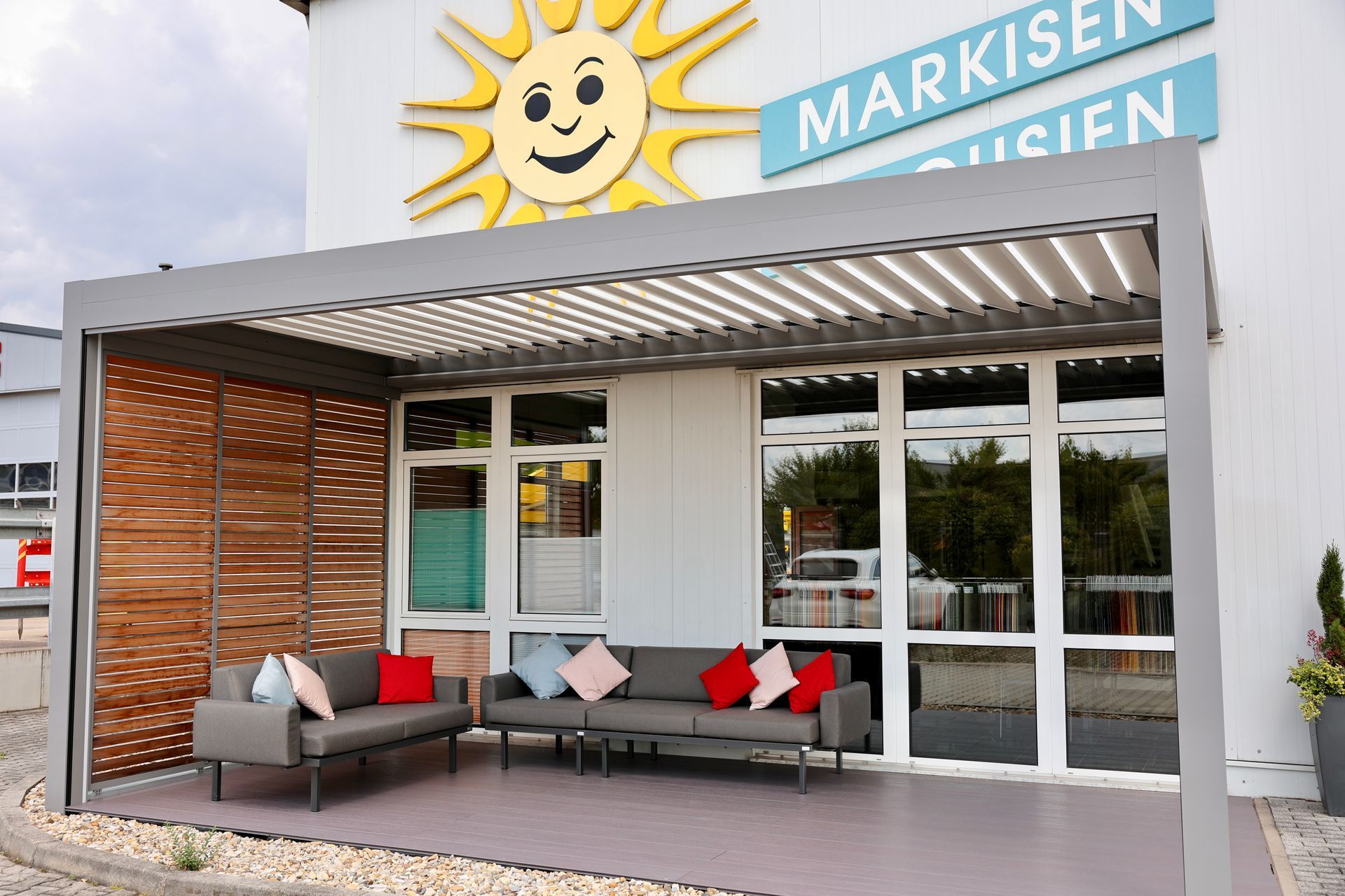 Ein moderner Sitzbereich im Freien unter einer Pergola. Graues Sofa mit roten und blauen Kissen. Ladenschild: „Markisen-Husien“.