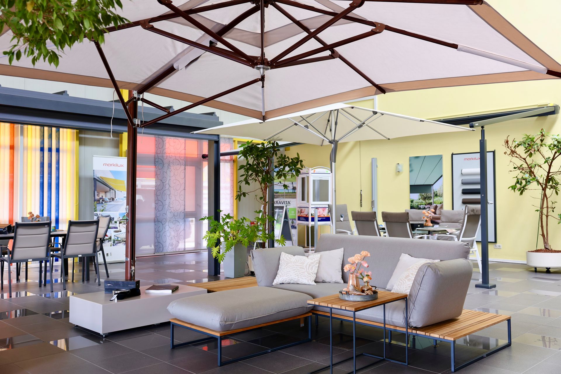 Gartenmöbelausstellung unter großen Sonnenschirmen. Bestehend aus einem grauen Sofa, Couchtischen und einer Essgruppe.